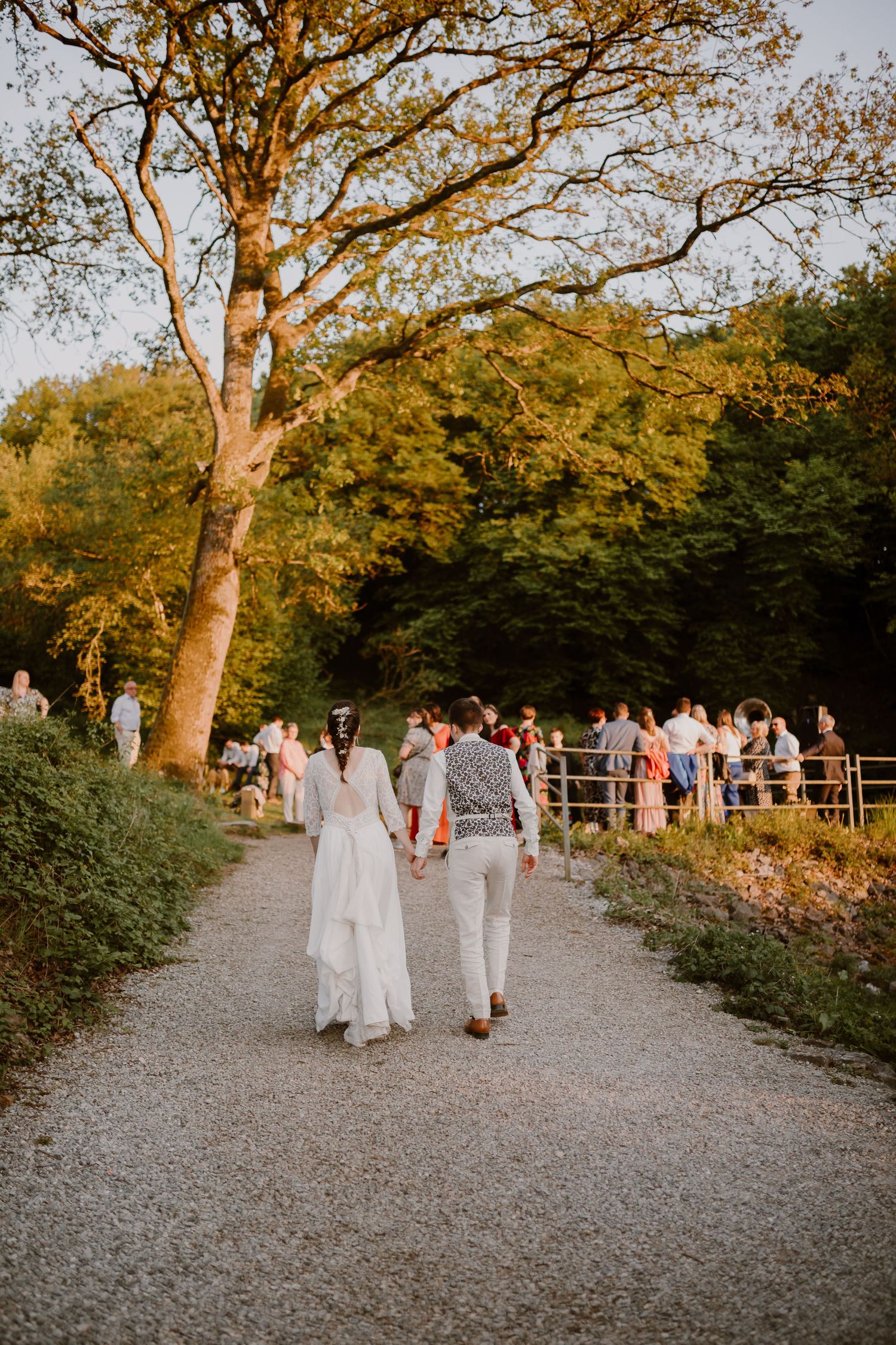 A couple in white outfits walks along a gravel path under autumn trees at sunset while guests gather in the background.