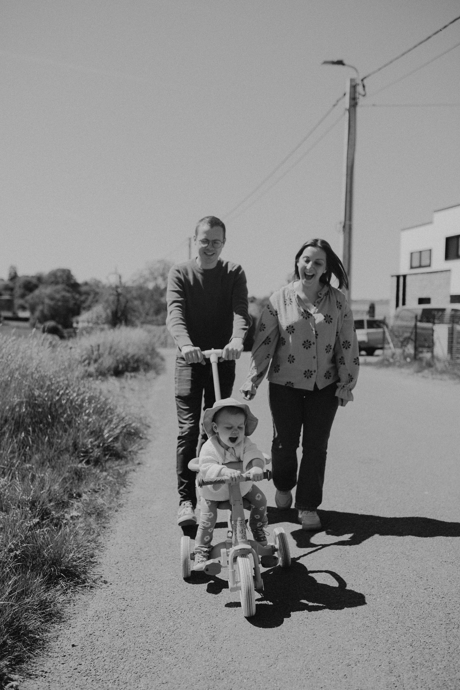 Black and white photo of family spending time together outdoors as child learns to ride a bicycle on sunny day.