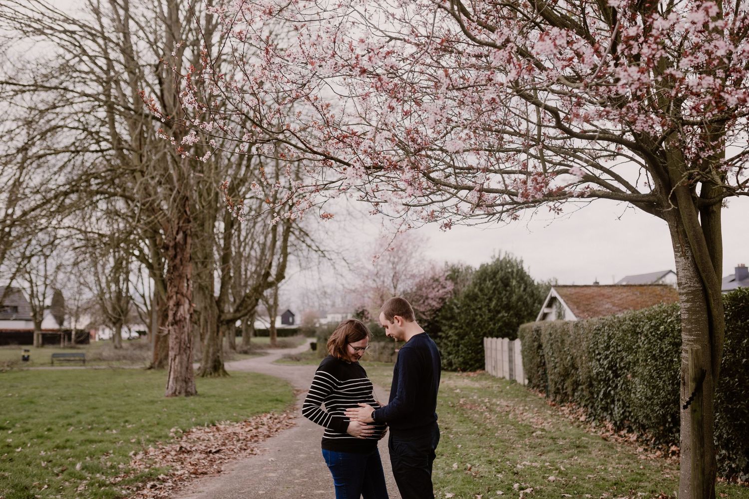 A couple stands on a path beneath pink cherry blossoms during an overcast spring day.