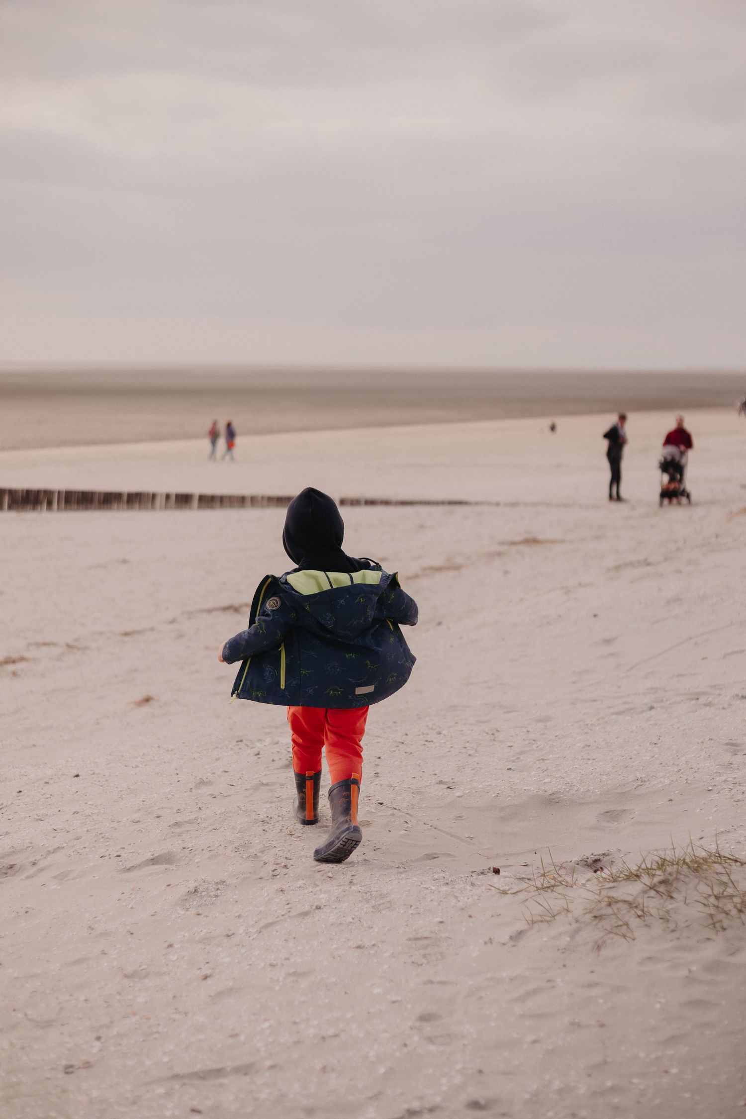 Person in red pants and navy jacket walking alone on a misty beach with other beachgoers visible in the distance.