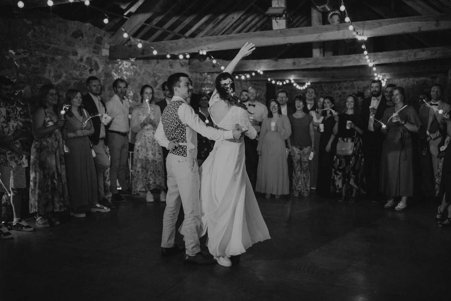 A couple shares their first dance at a rustic barn wedding reception with string lights and guests looking on.