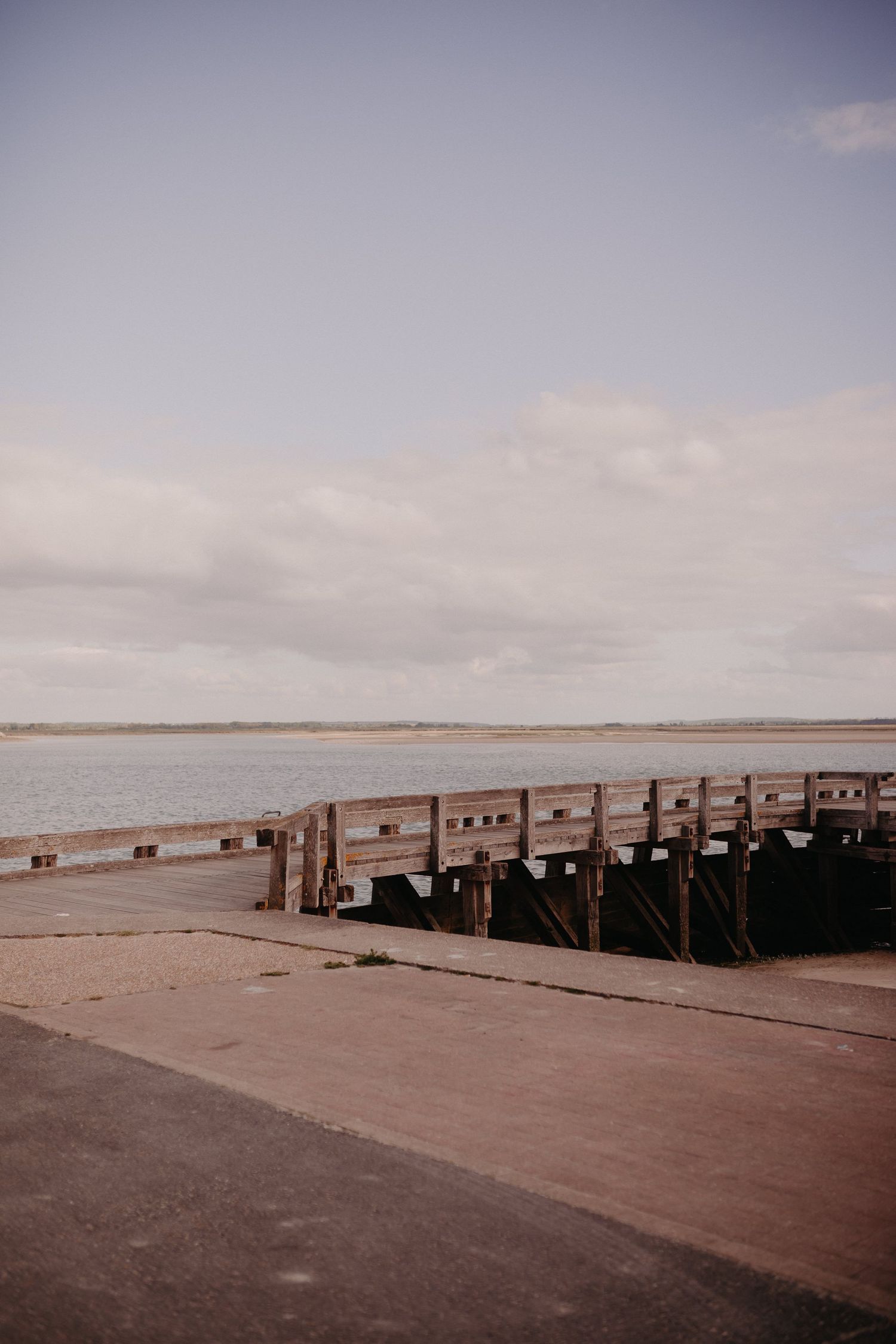 A wooden pier extends into a calm lake under an overcast sky with a concrete pathway in the foreground.