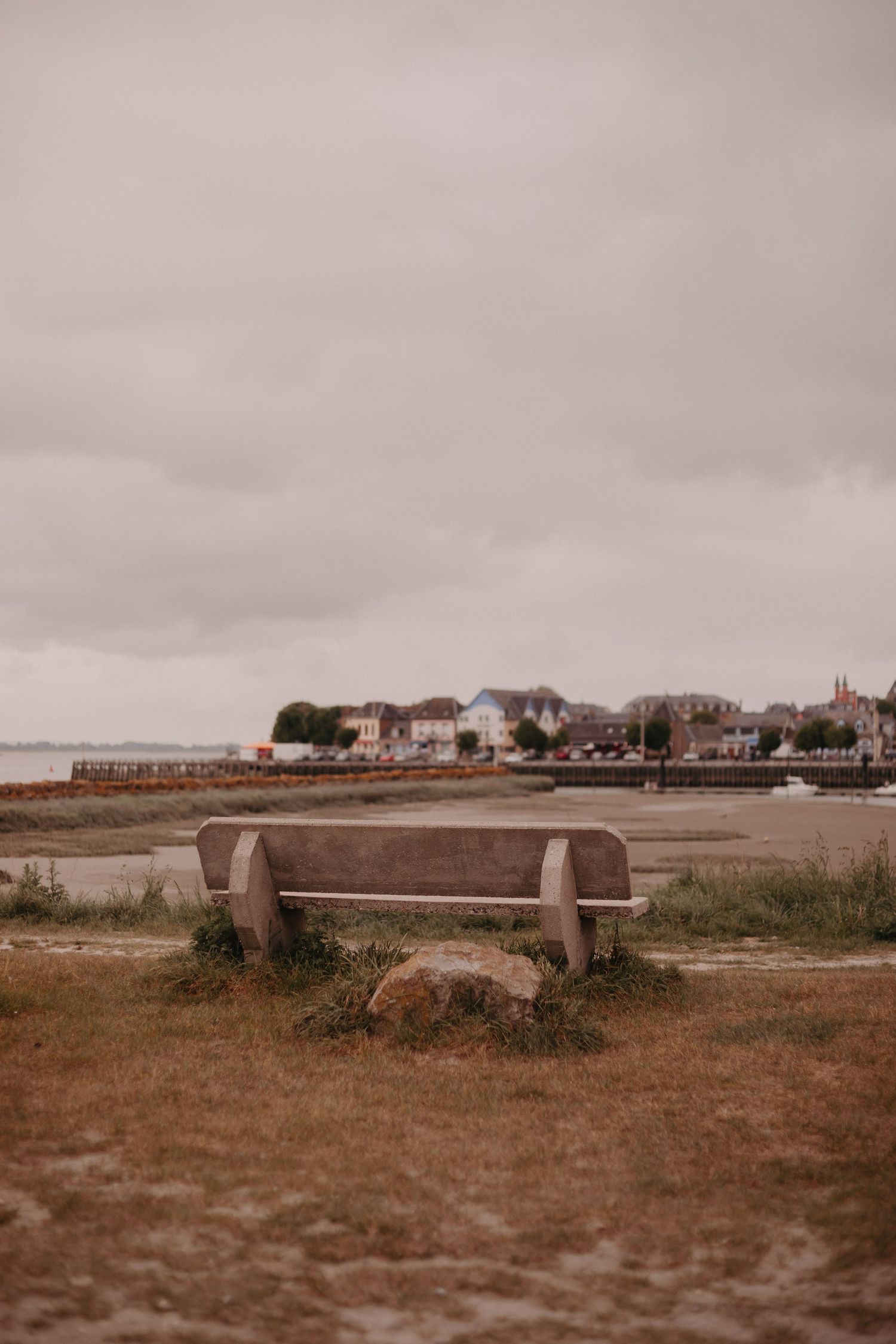 Empty wooden bench overlooking a coastal town with houses and a cloudy sky in muted, vintage tones.