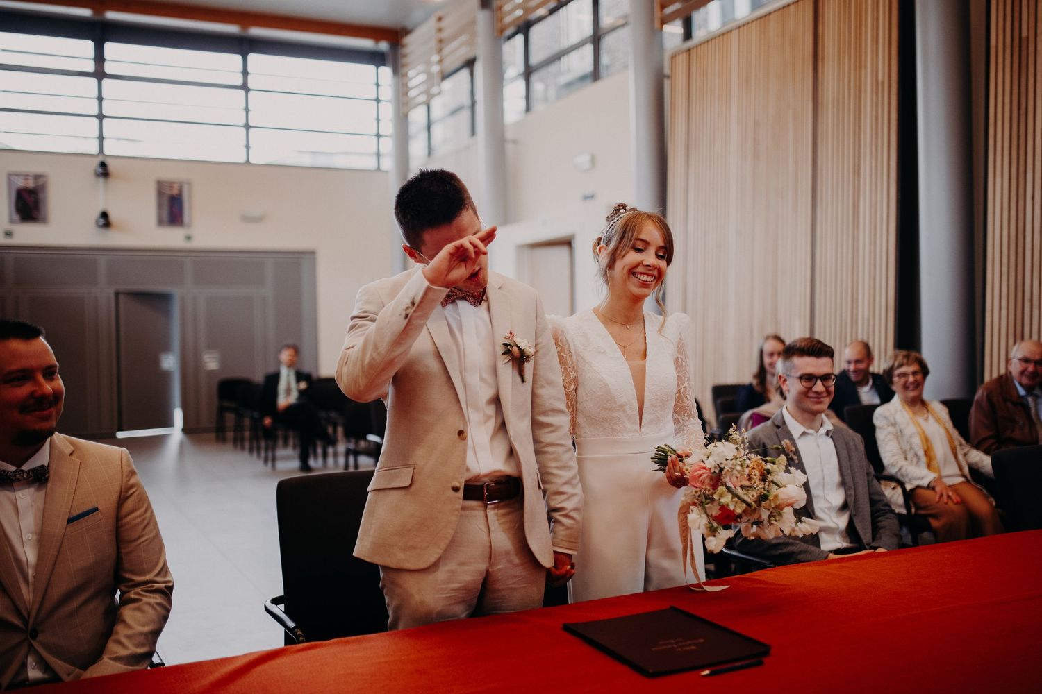 Wedding couple laughing during indoor ceremony in front of brick wall with guests seated to the side.