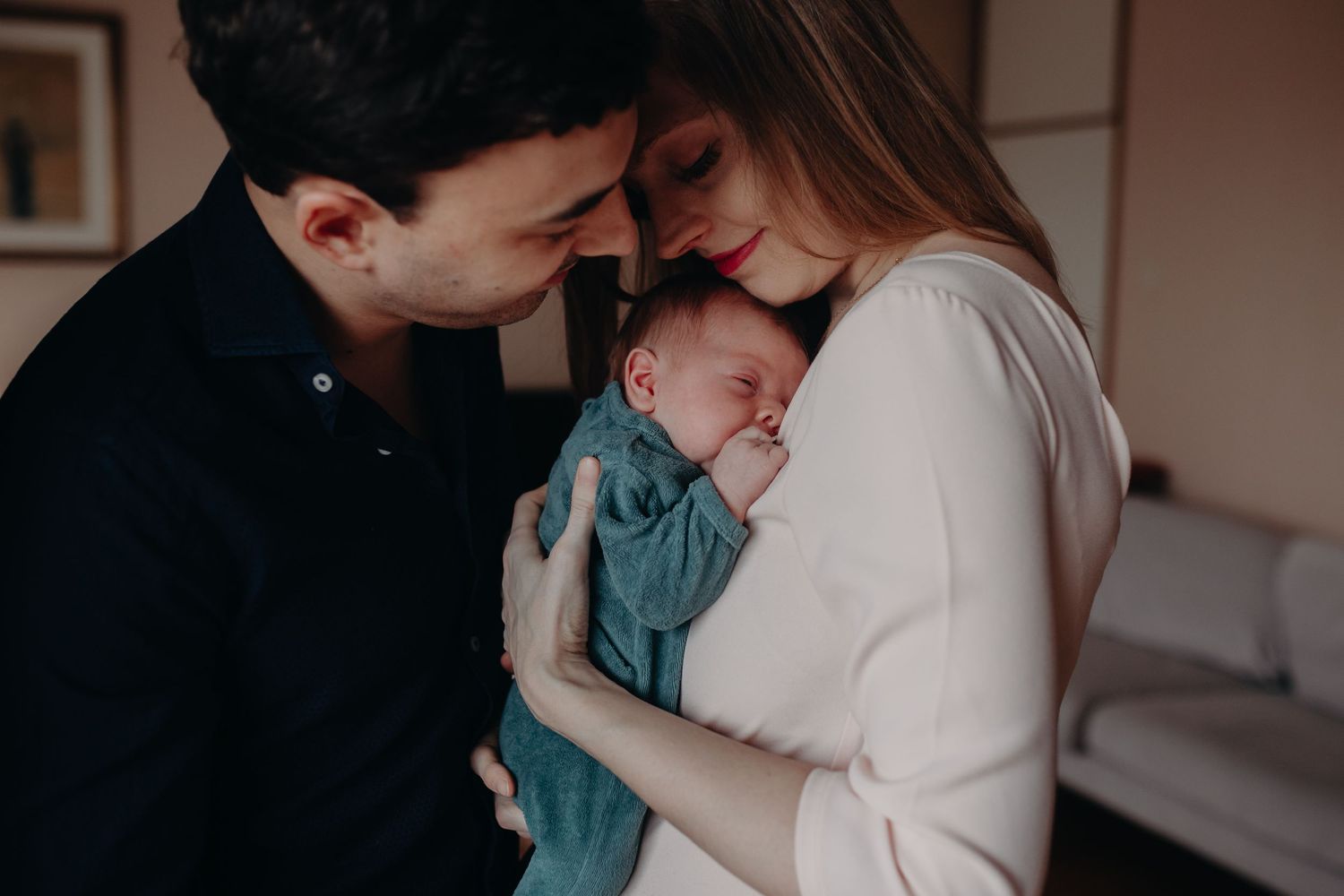 A tender family moment as two people embrace their sleeping newborn baby wrapped in a teal swaddle.