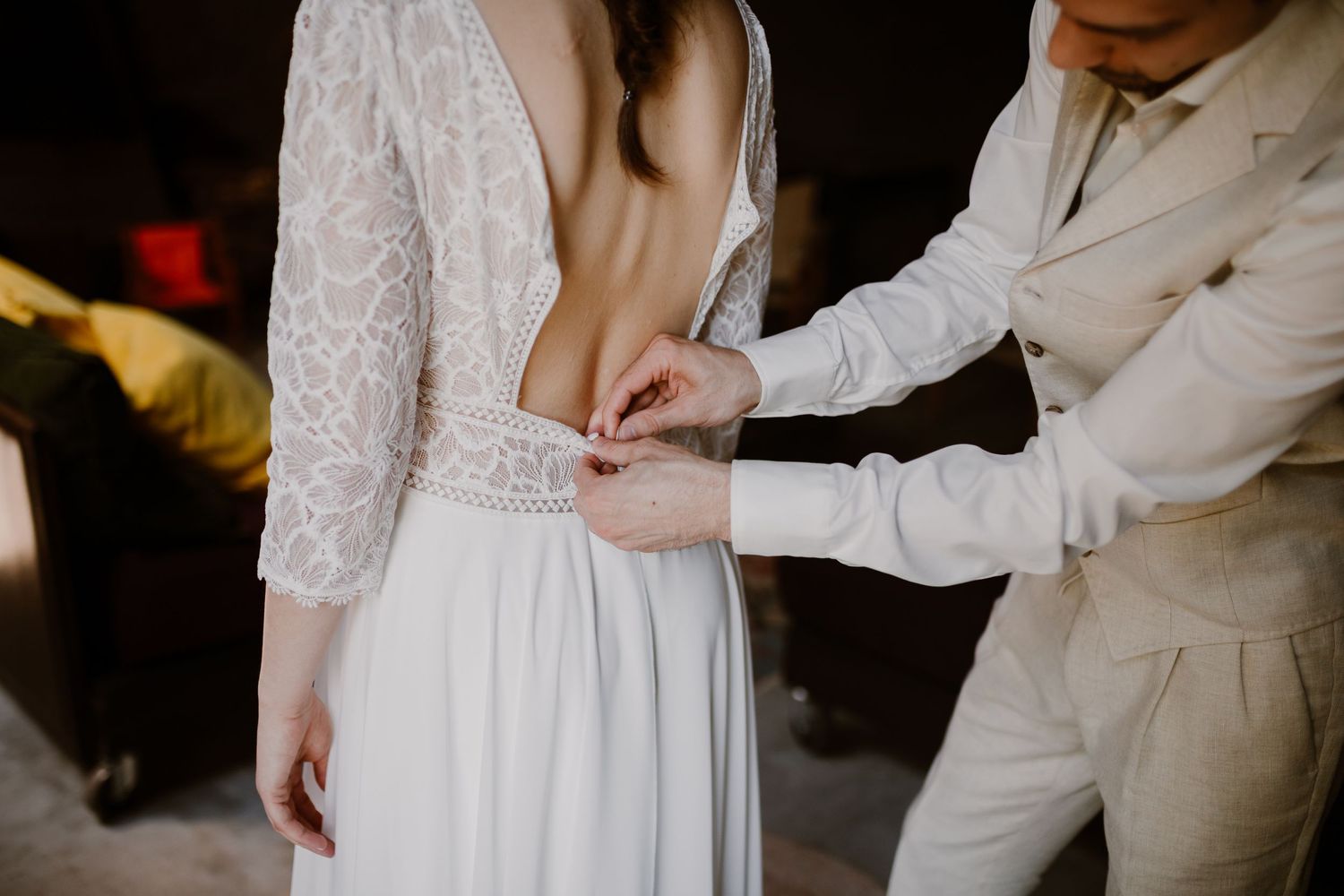 Close up of a white lace wedding dress being buttoned up in the back by someone wearing a light colored suit.