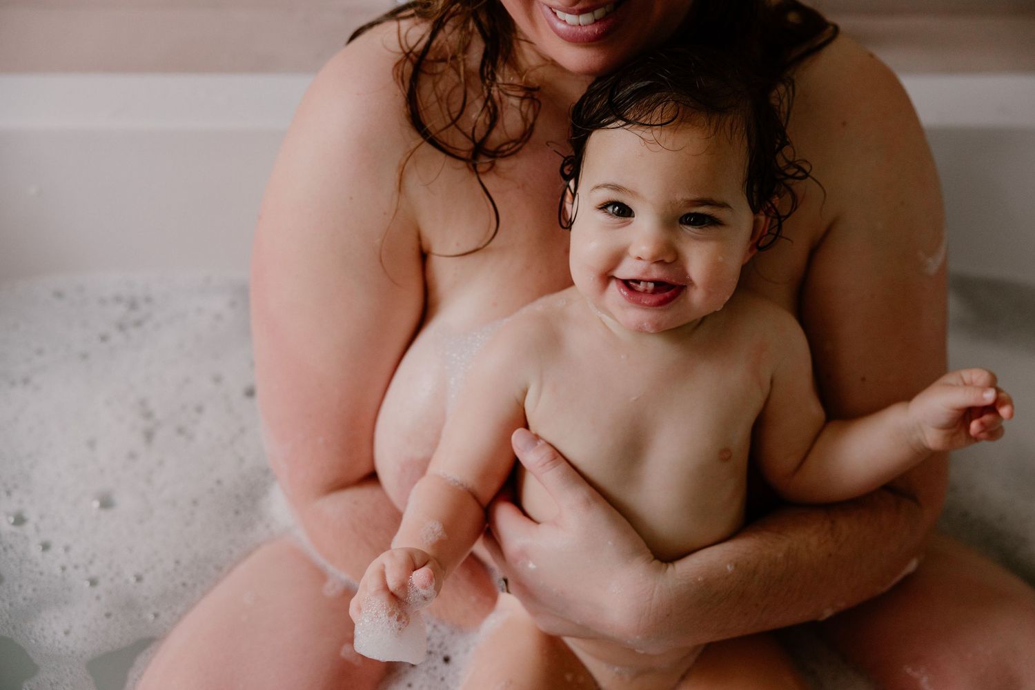 Mother and baby sharing a joyful bubble bath together, smiling infant sitting in parent's arms.