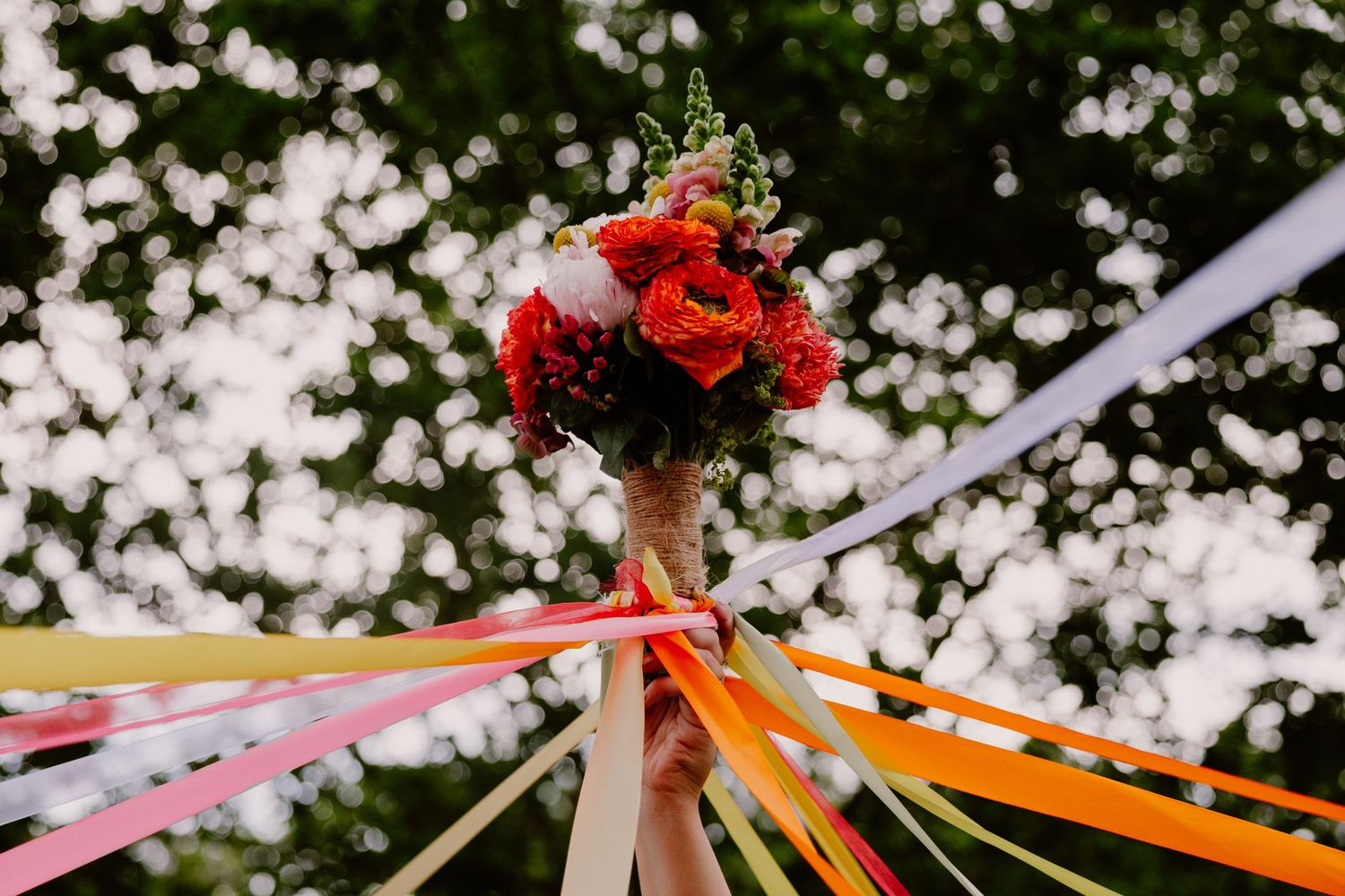 Hands holding a vibrant red floral bouquet with colorful streaming ribbons outdoors at a wedding ceremony.