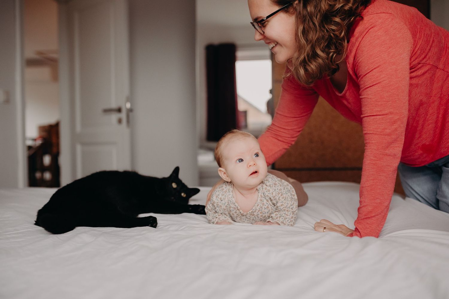 A baby lies on a bed next to a black cat while someone in a red shirt leans over them.