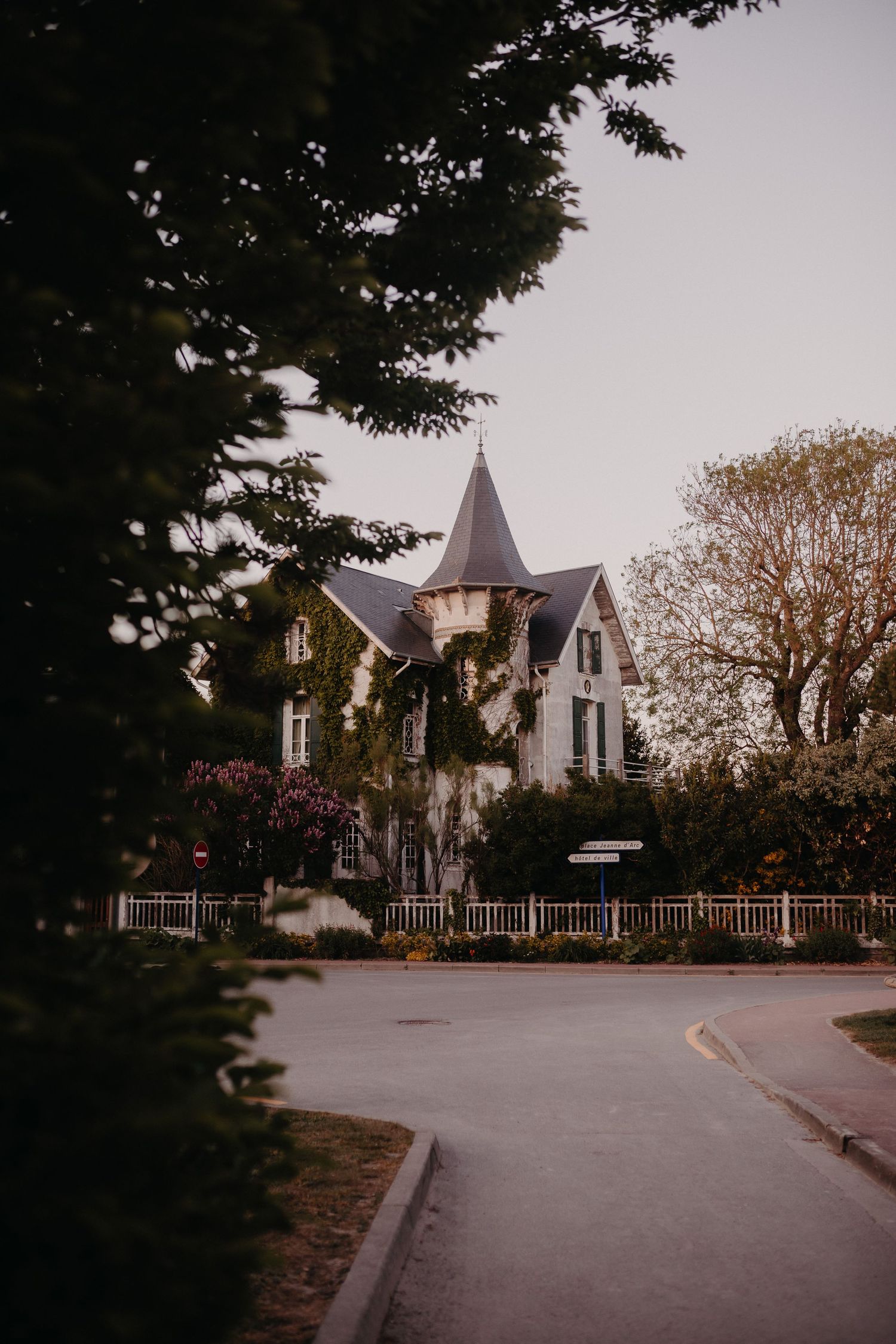 A charming Victorian house with a pointed tower stands behind a white picket fence on a curved residential street.
