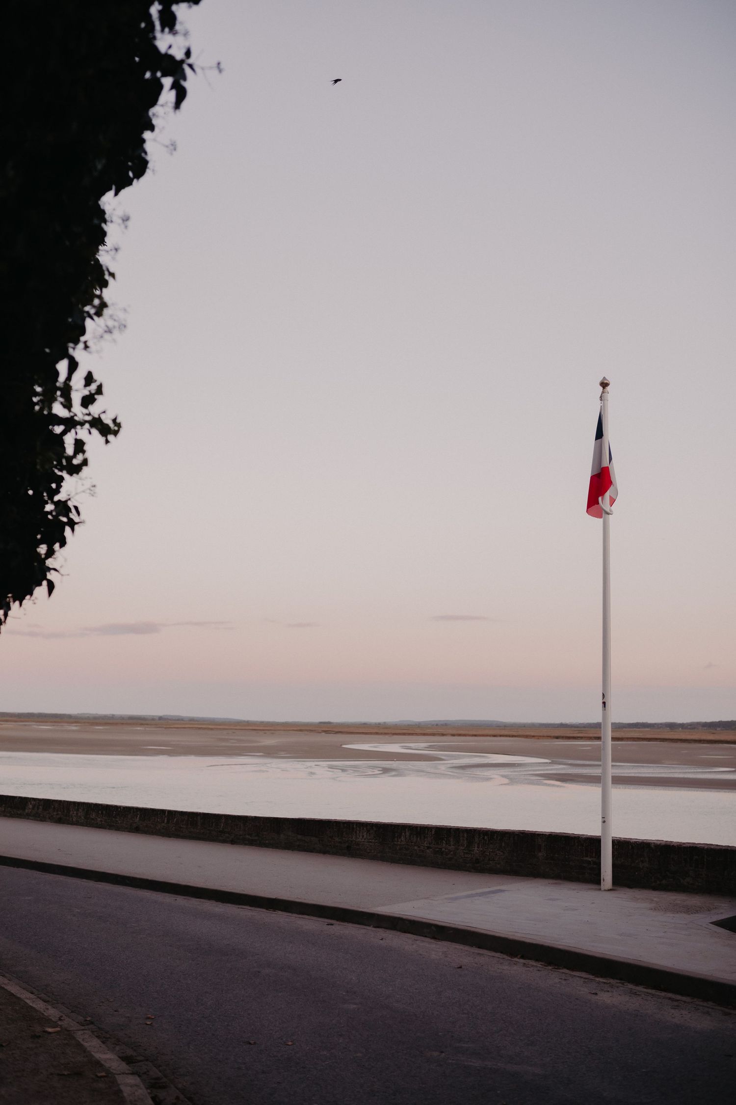 A French flag waves on a pole along an empty coastal road at dusk with a beach and calm waters in the background.