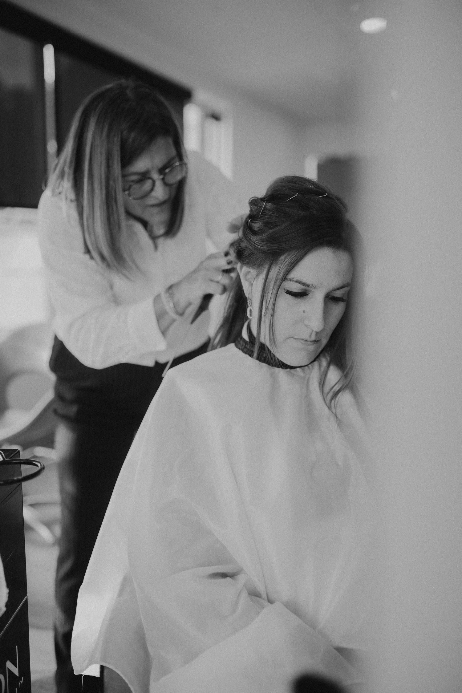 Black and white photo of a hairstylist styling bride's hair during wedding preparations in an intimate indoor setting.