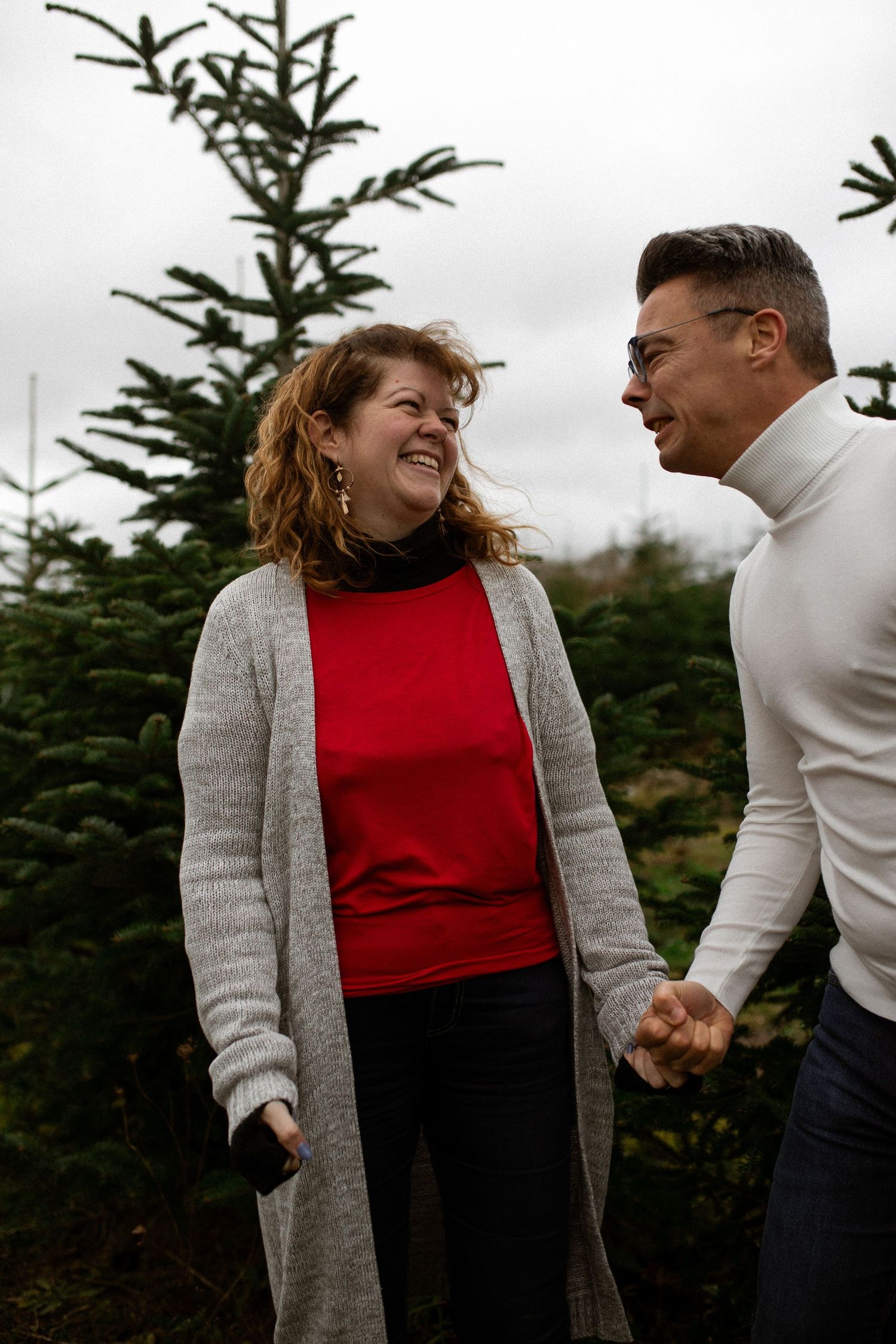 A couple in winter attire shares a joyful moment together among evergreen trees on an overcast day.