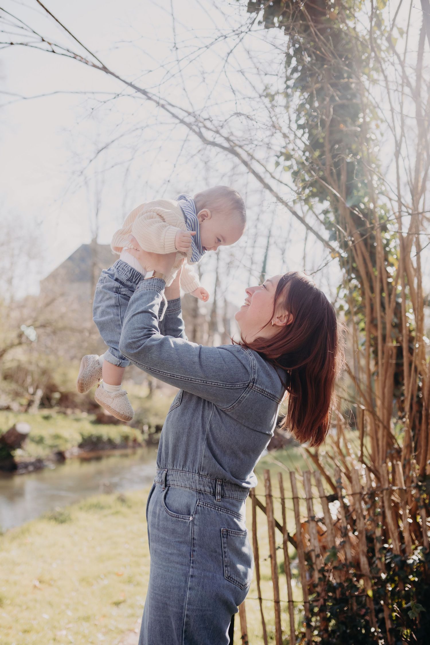 A person in denim overalls lifts a small child up against a natural outdoor backdrop with trees and water.