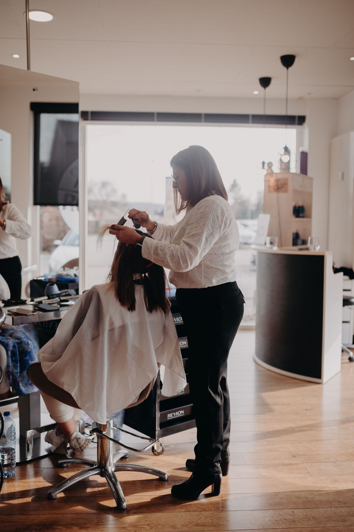 A hairstylist in a white shirt and black pants works on a client's hair at a modern salon with hardwood floors.