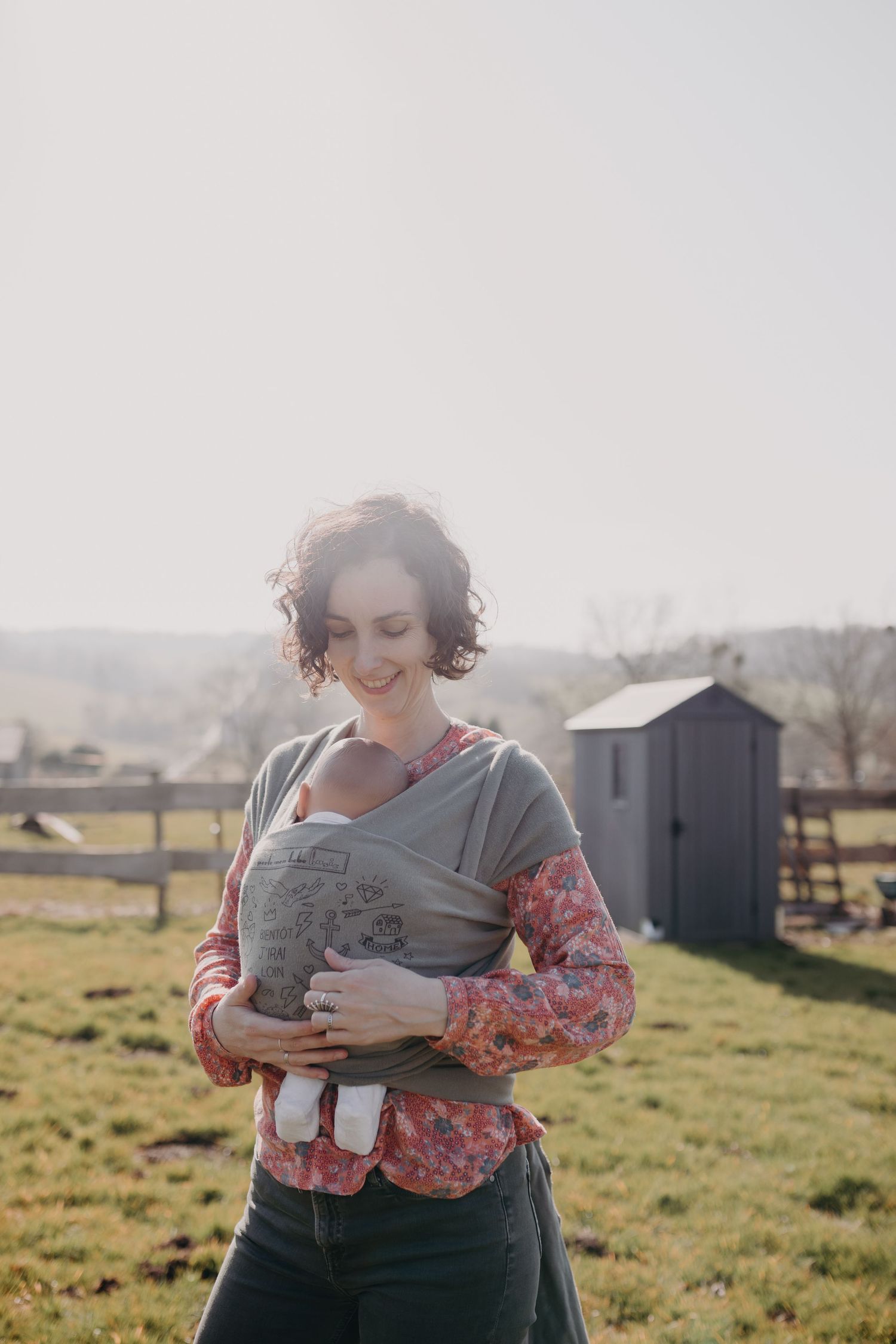 Person wearing a gray baby carrier and floral top stands in a sunlit rural field with a barn in the background.
