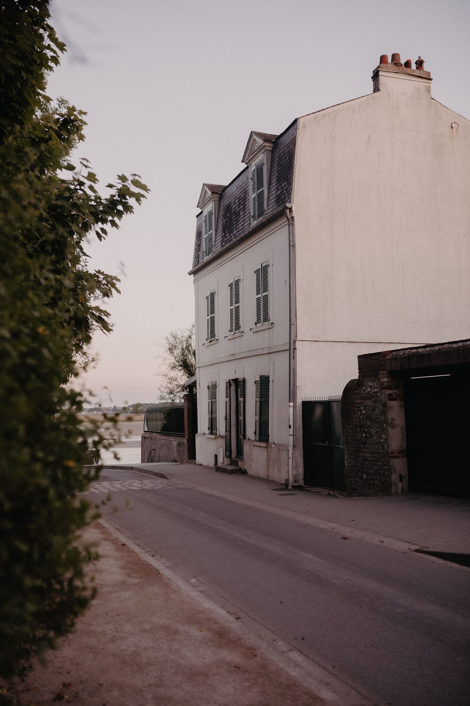 A white three-story townhouse with dormer windows stands along a curved street in an old European neighborhood.