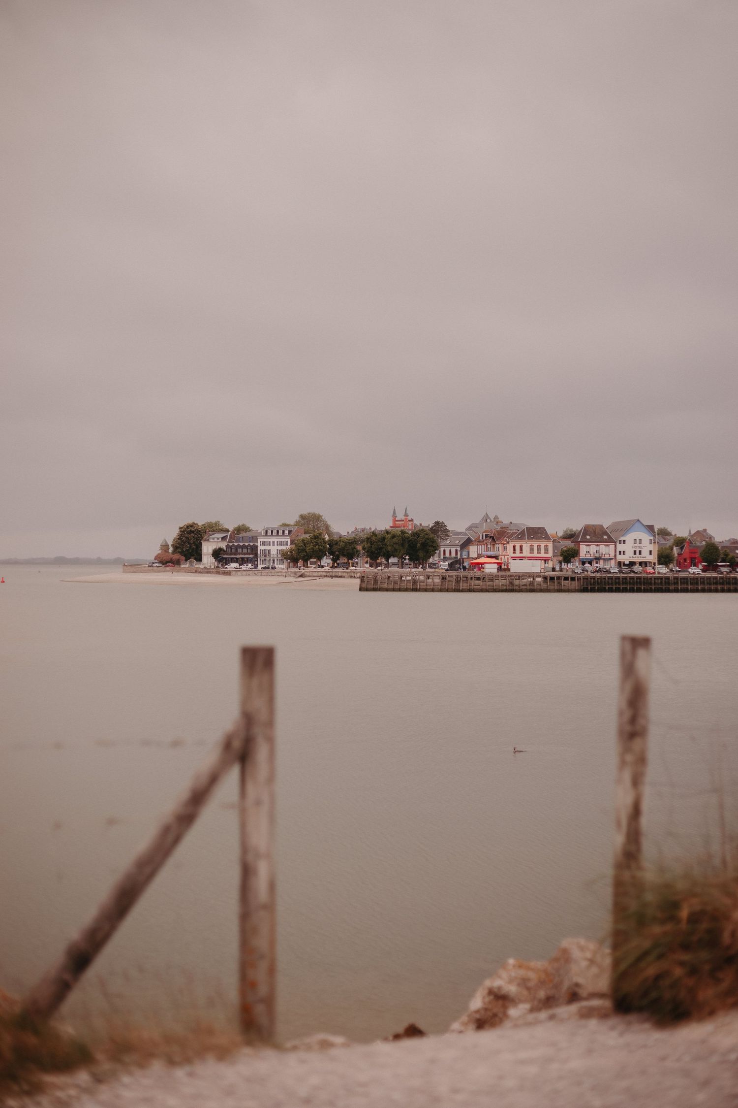 Coastal houses line the waterfront on a misty day, viewed through wooden fence posts along the shoreline.