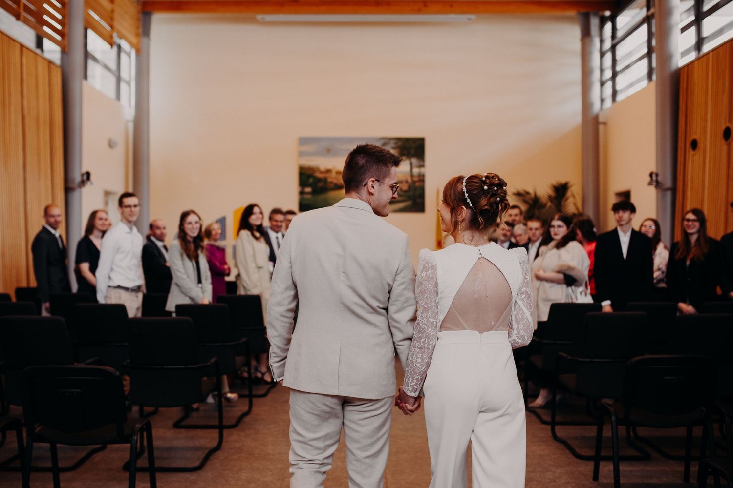 A couple holds hands at a small indoor ceremony with guests seated in rows of black chairs.