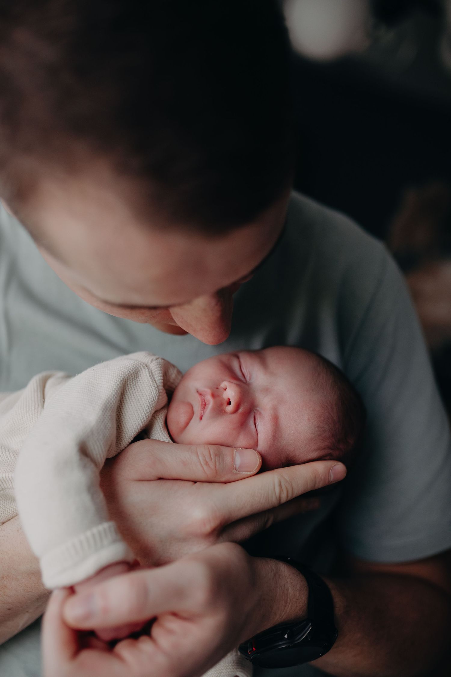 A sleeping newborn baby swaddled in white blankets being held against someone wearing gray.