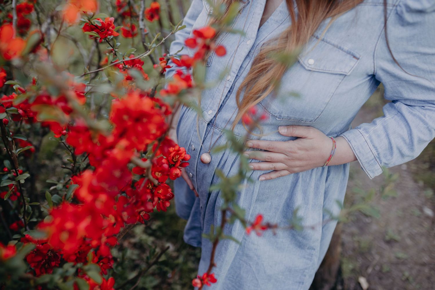 A pregnant woman in a denim jacket stands next to vibrant red flowering bushes in a garden setting.