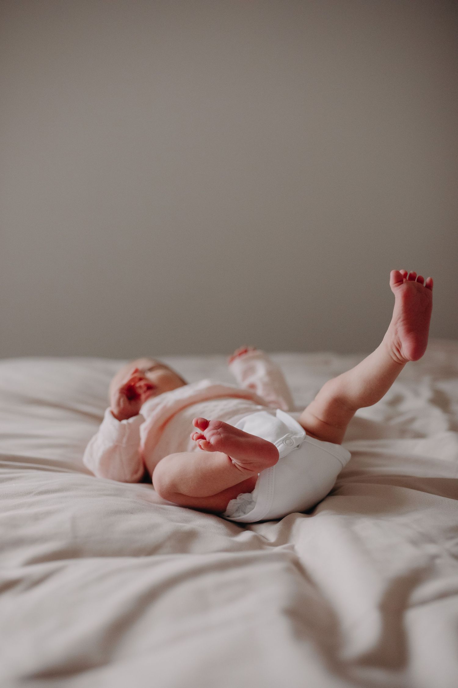 Baby's tiny feet and legs peek out from white fabric while lying on soft beige bedding.