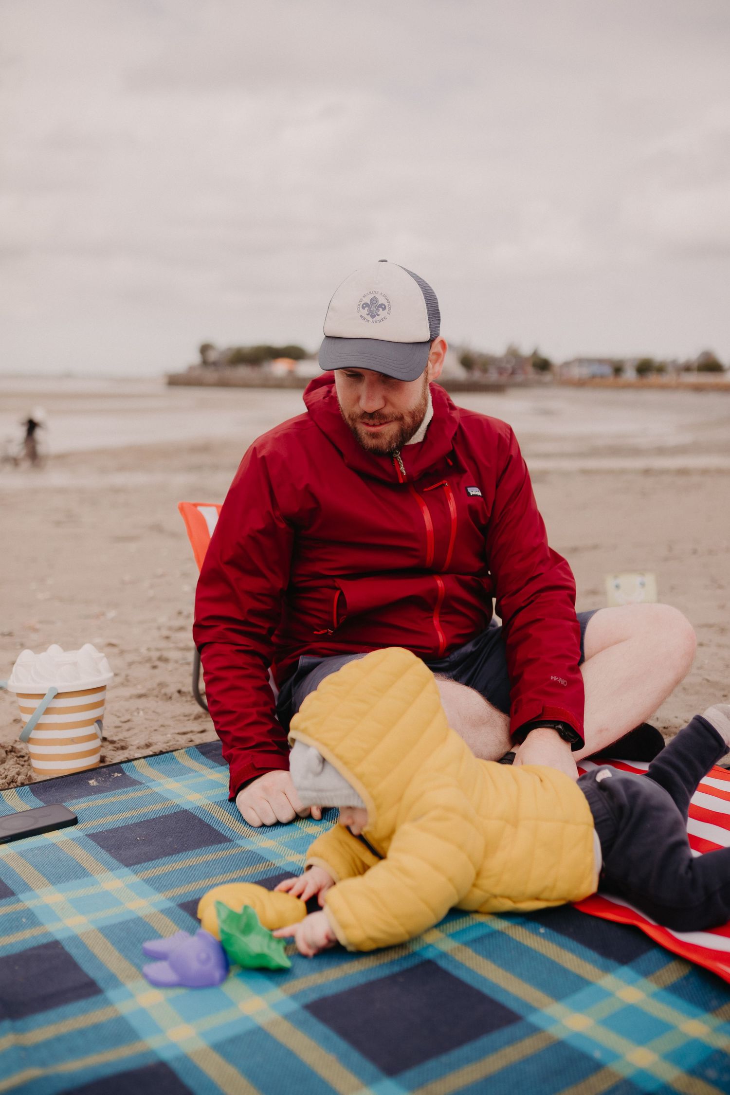 A person in a red jacket sits on a plaid blanket at the beach with a child in a yellow snowsuit playing with toys.