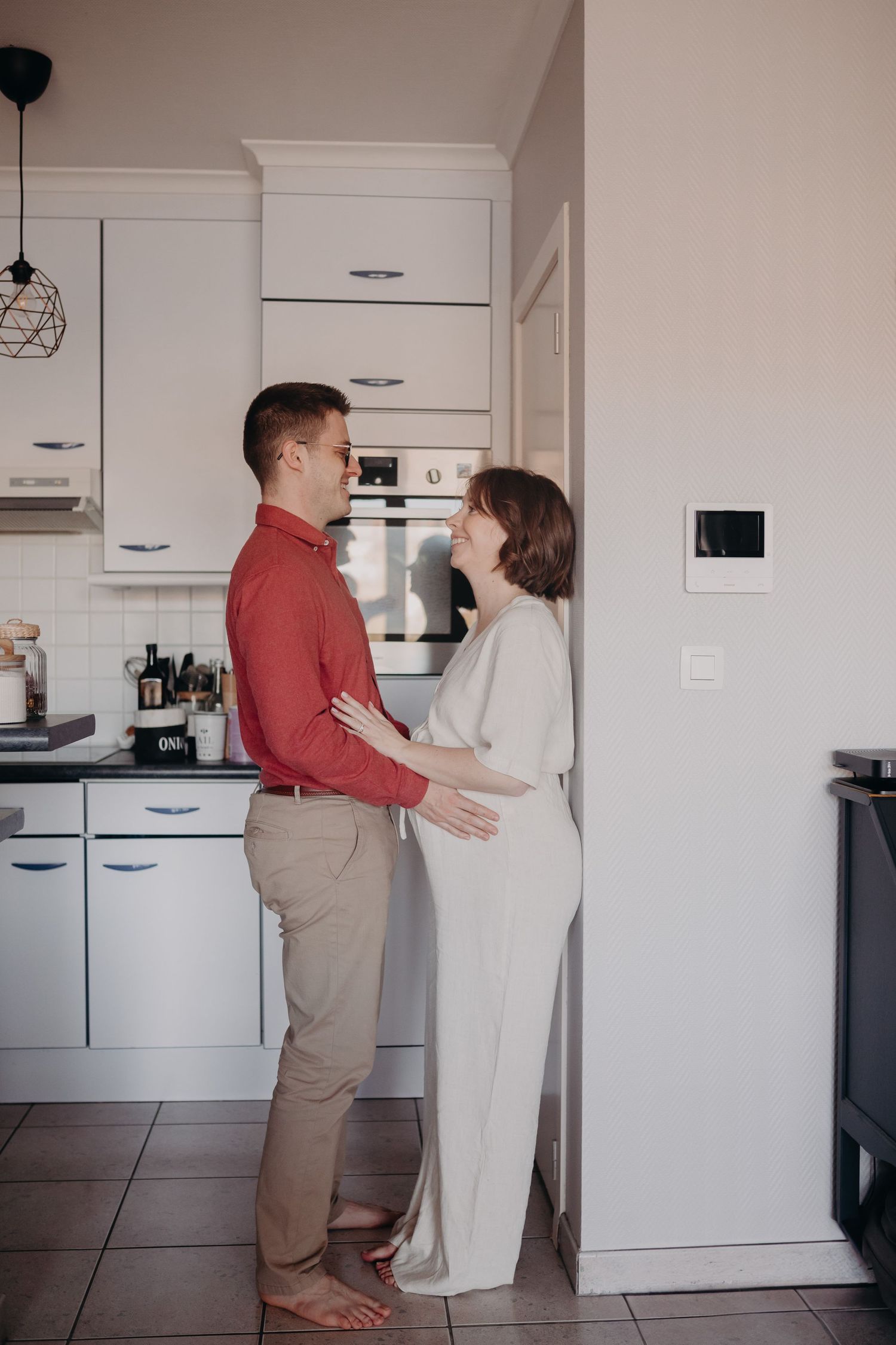 A couple stands lovingly together in a modern white kitchen with pendant lighting and tiled floors.