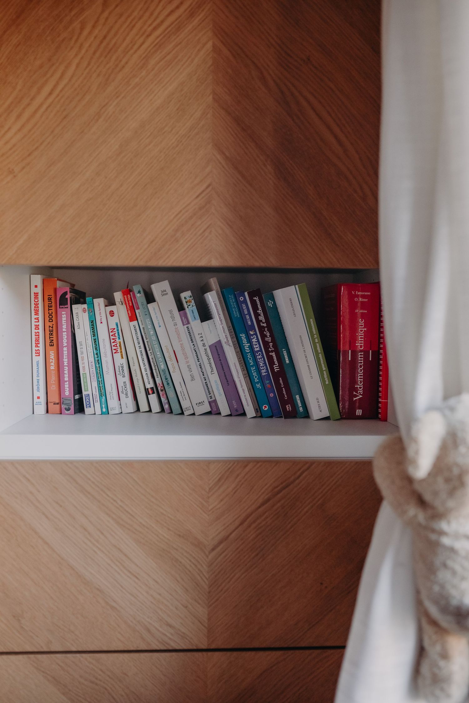 Colorful books neatly arranged on a wooden shelf next to a white curtain in a minimalist setting.