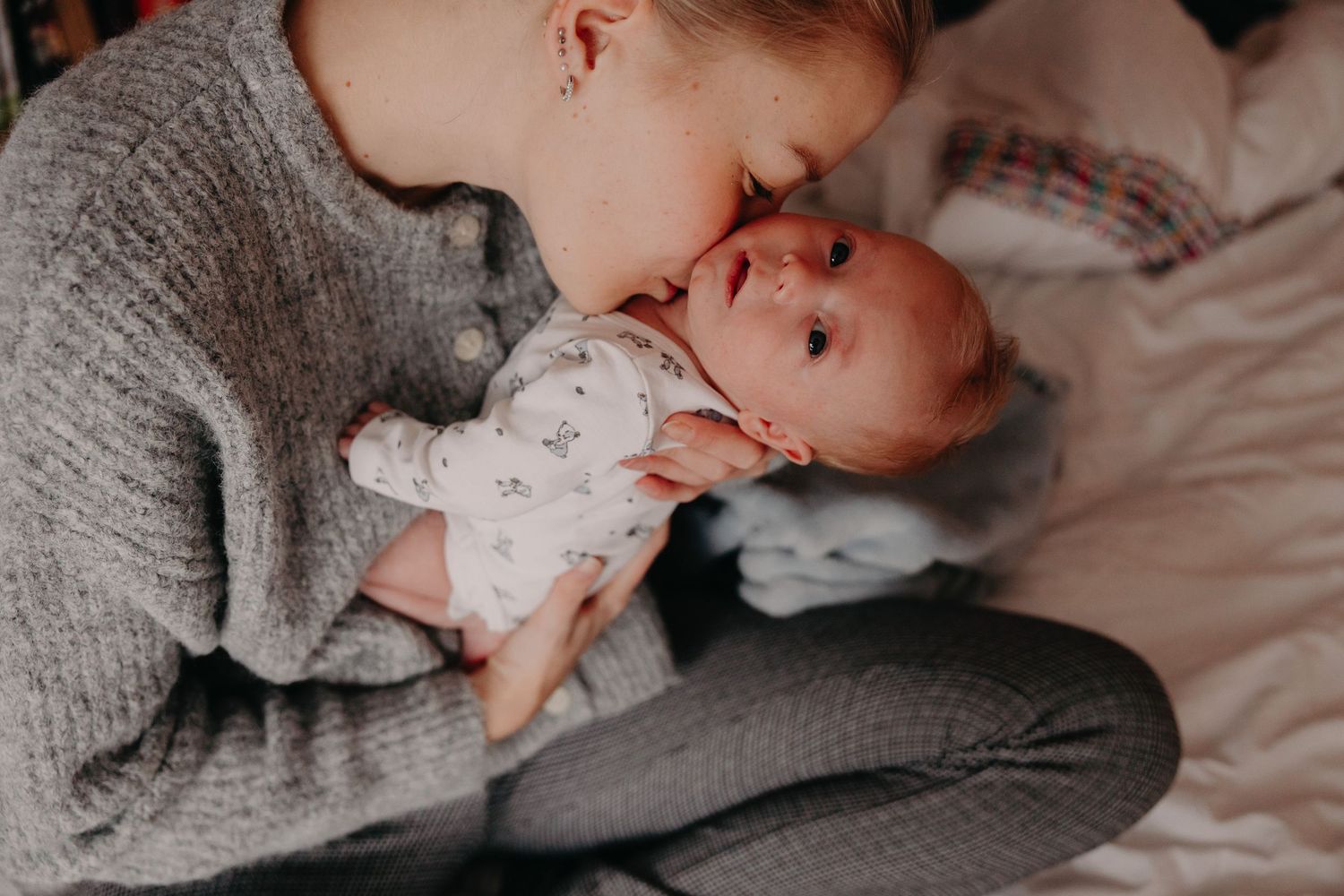 A tender moment between parent and baby lying on white bedding while wearing cozy gray sweaters.