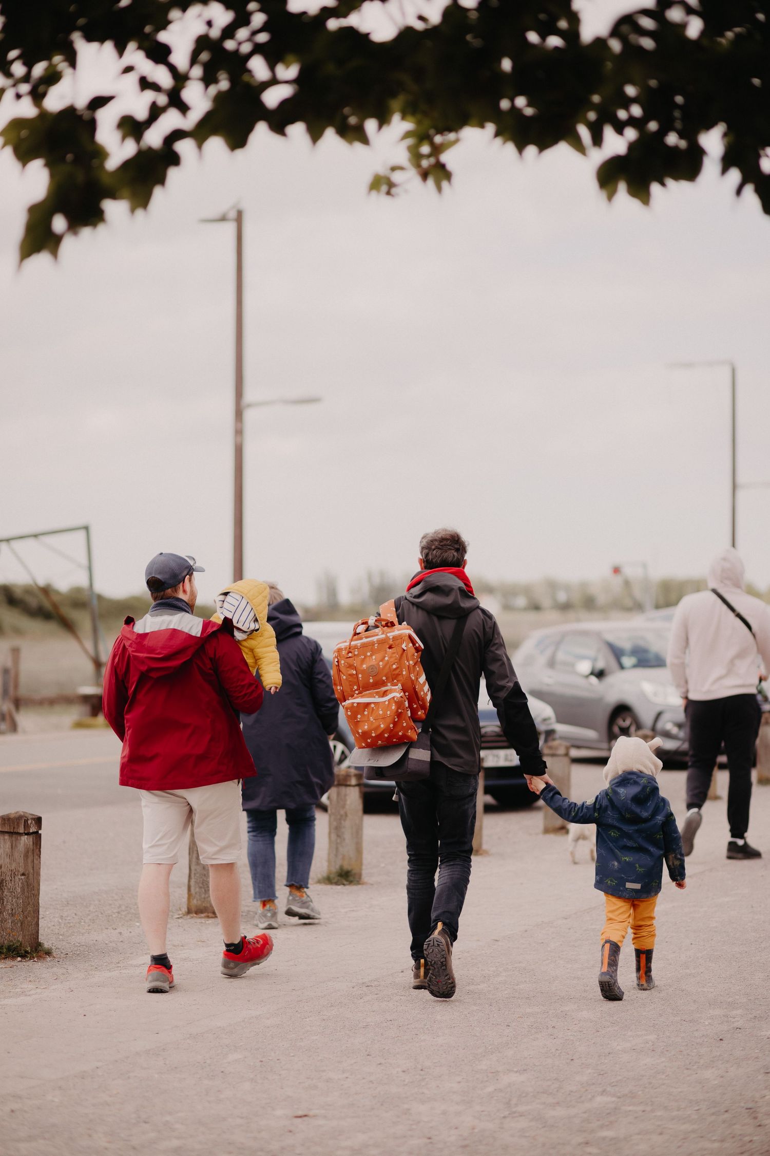 People walk together on a path near parked cars on an overcast day, with tree branches visible overhead.