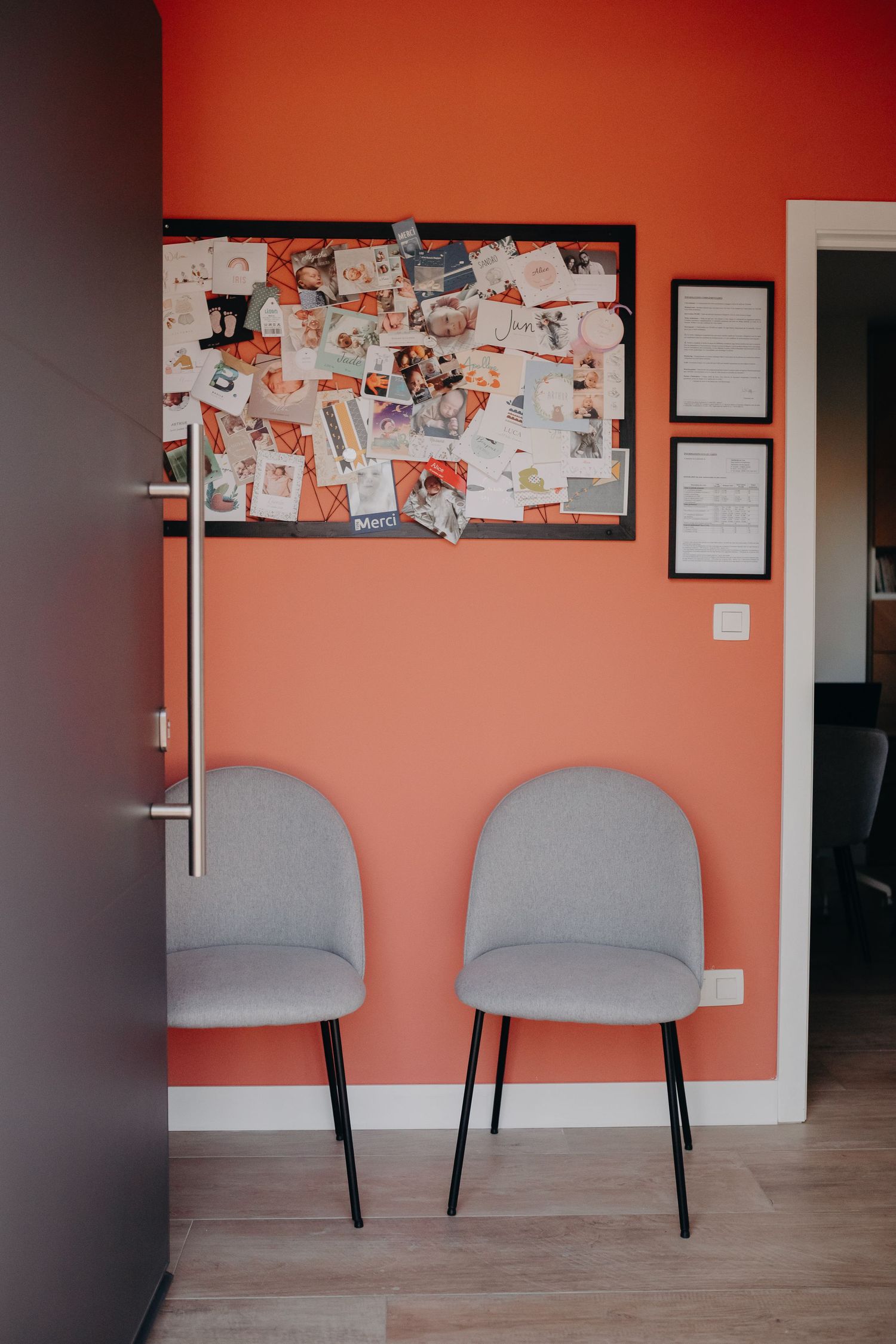 Two gray upholstered dining chairs sit against a coral-colored wall with a bulletin board displaying photos and papers.