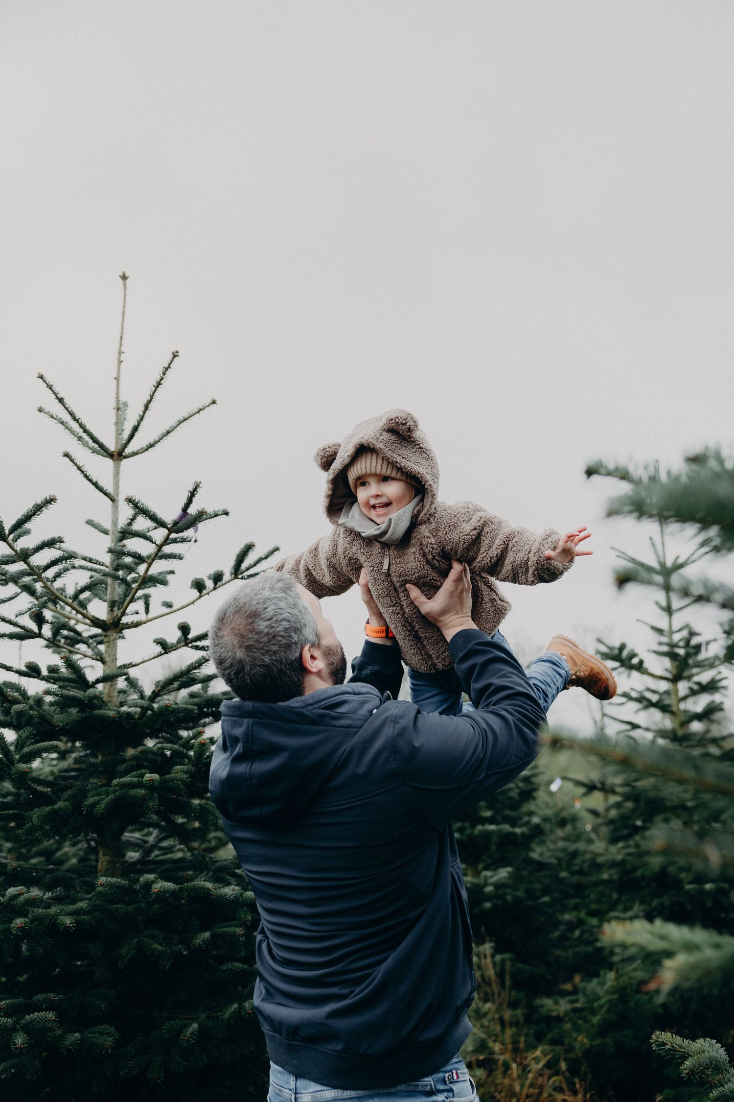 A person lifts up a child while standing among evergreen trees at a Christmas tree farm on a cloudy day.