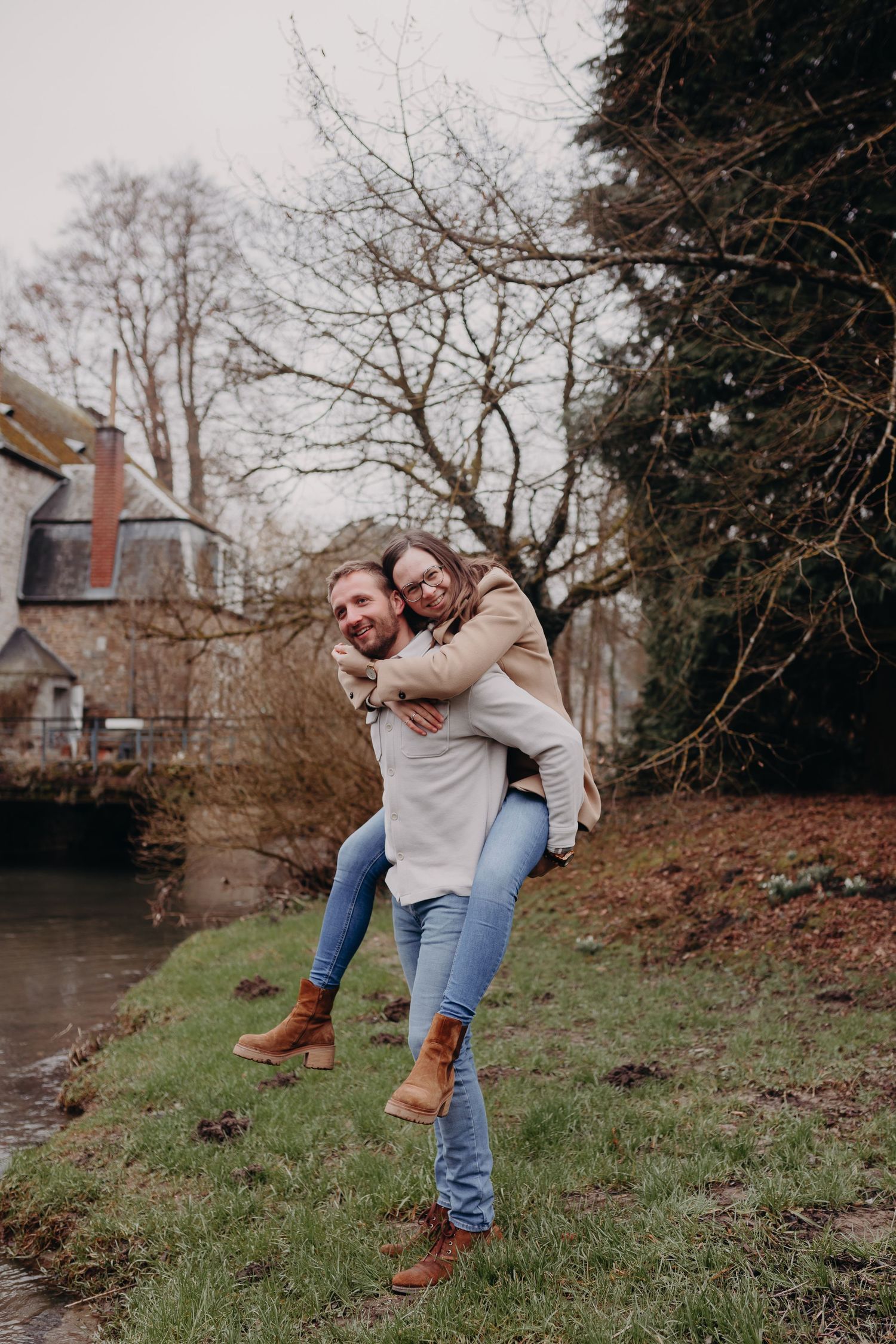 A playful piggyback ride moment captured on a grassy riverbank with bare trees and a building in the background.