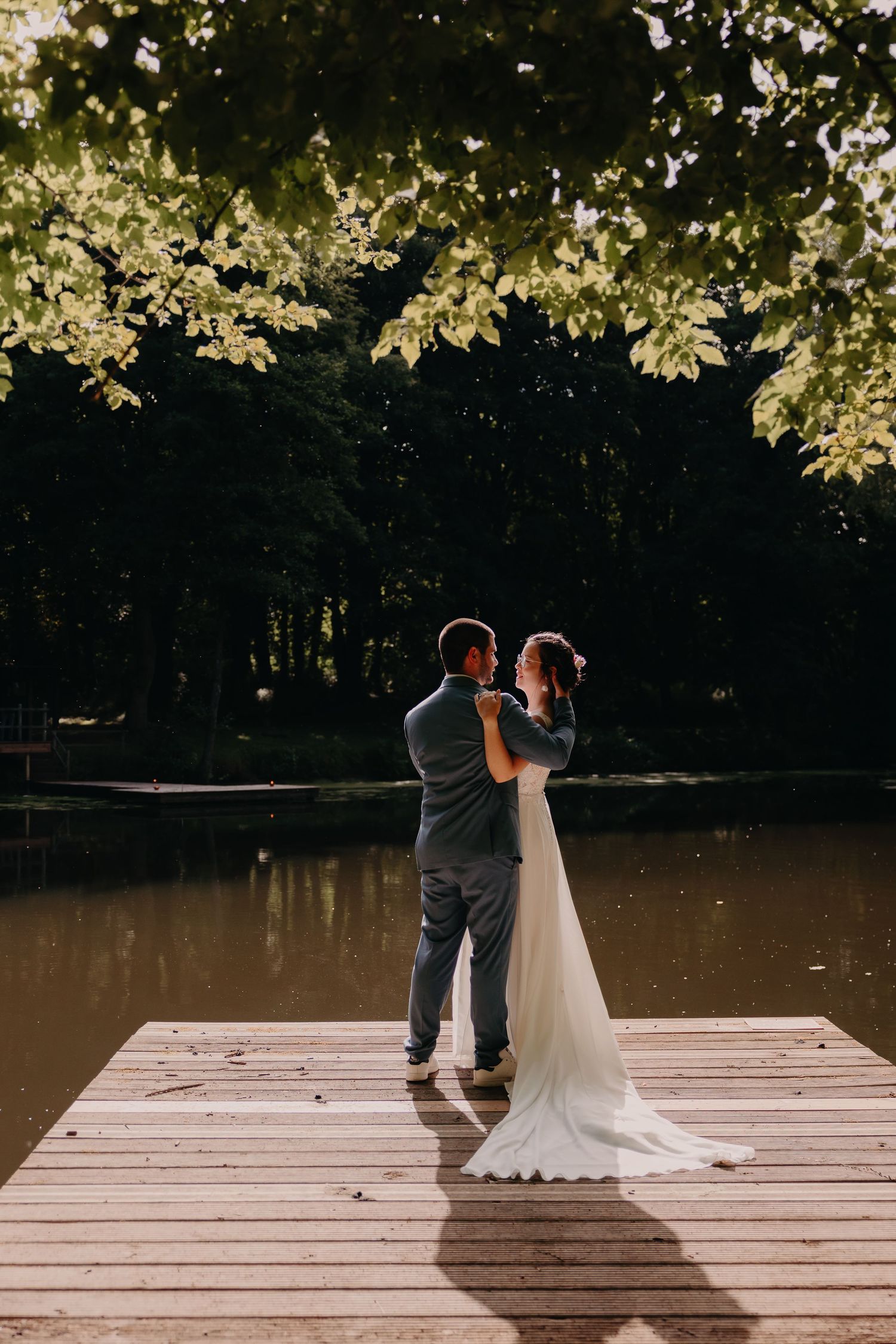 Newlyweds share an intimate moment on a wooden dock surrounded by overhanging trees at sunset.