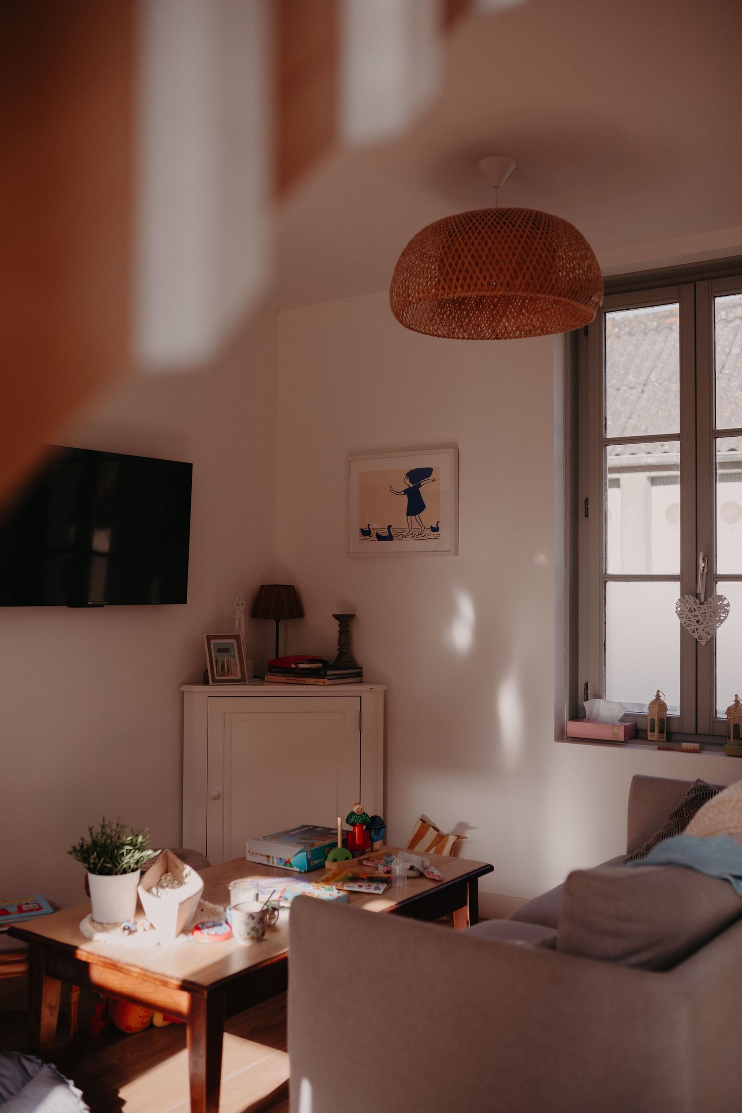 Cozy living room interior with rattan pendant lamp, wall-mounted TV, and soft gray sofa in warm lighting.
