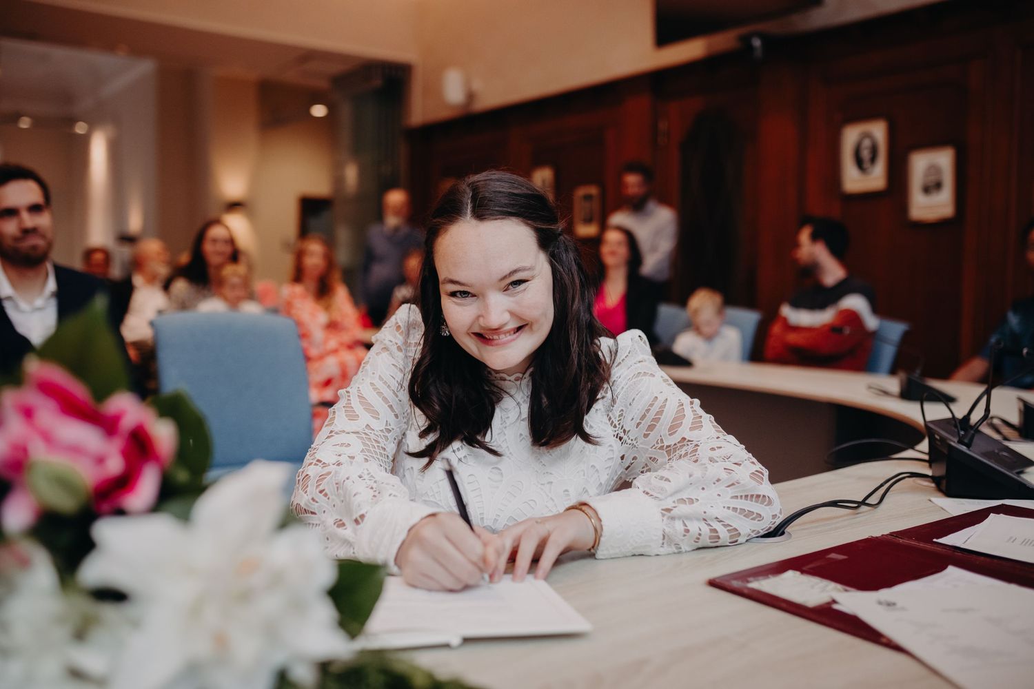 Someone signs paperwork at desk during celebratory event with guests and floral decorations in background.