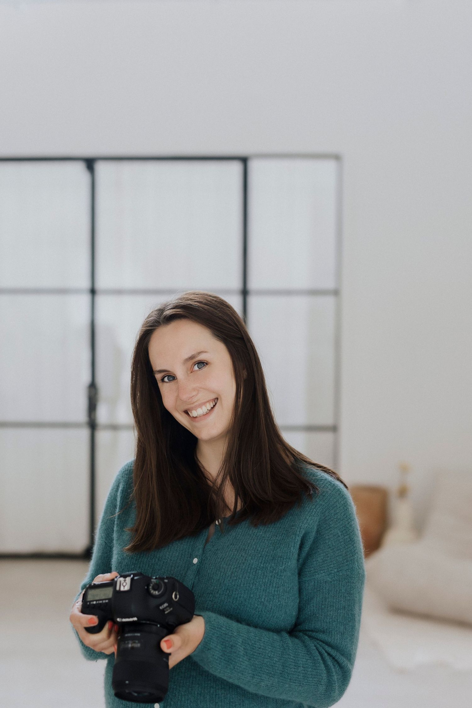 Professional photographer in a green sweater holds a DSLR camera while smiling in a bright indoor setting.