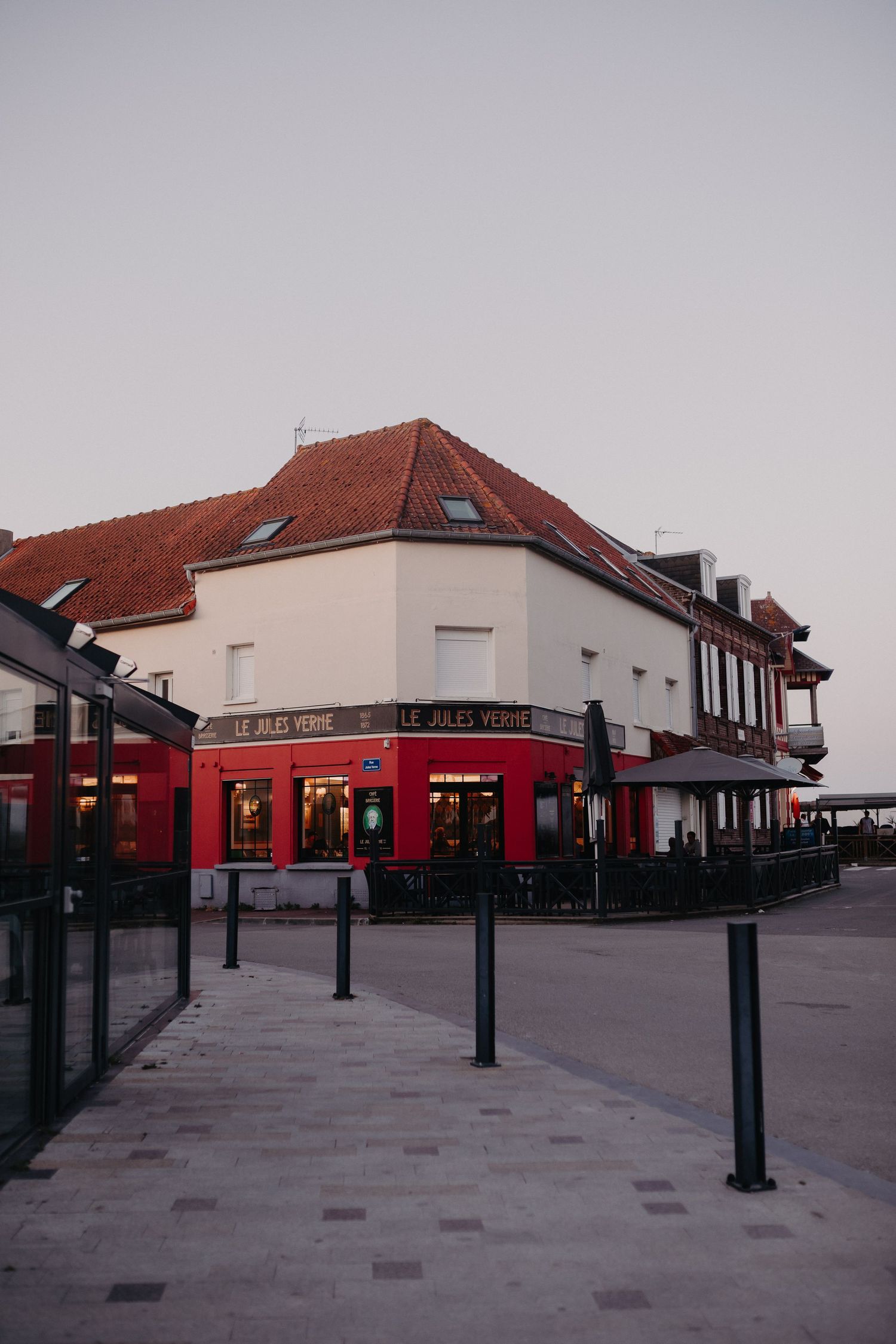 A corner restaurant building with red awnings and white walls at dusk in a European-style plaza.