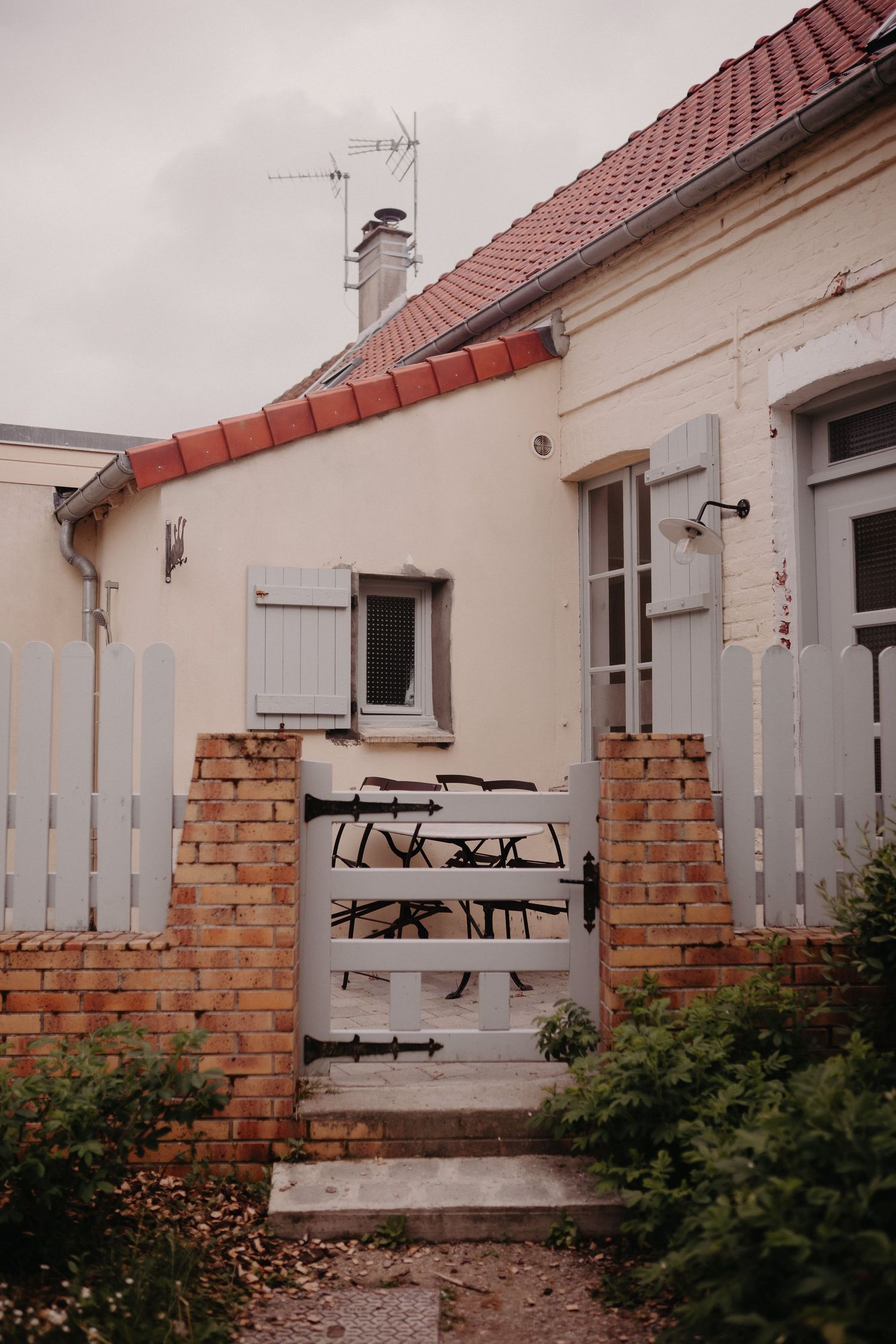 A rustic white house with red tile roof and brick fence entrance featuring a metal gate and worn steps.