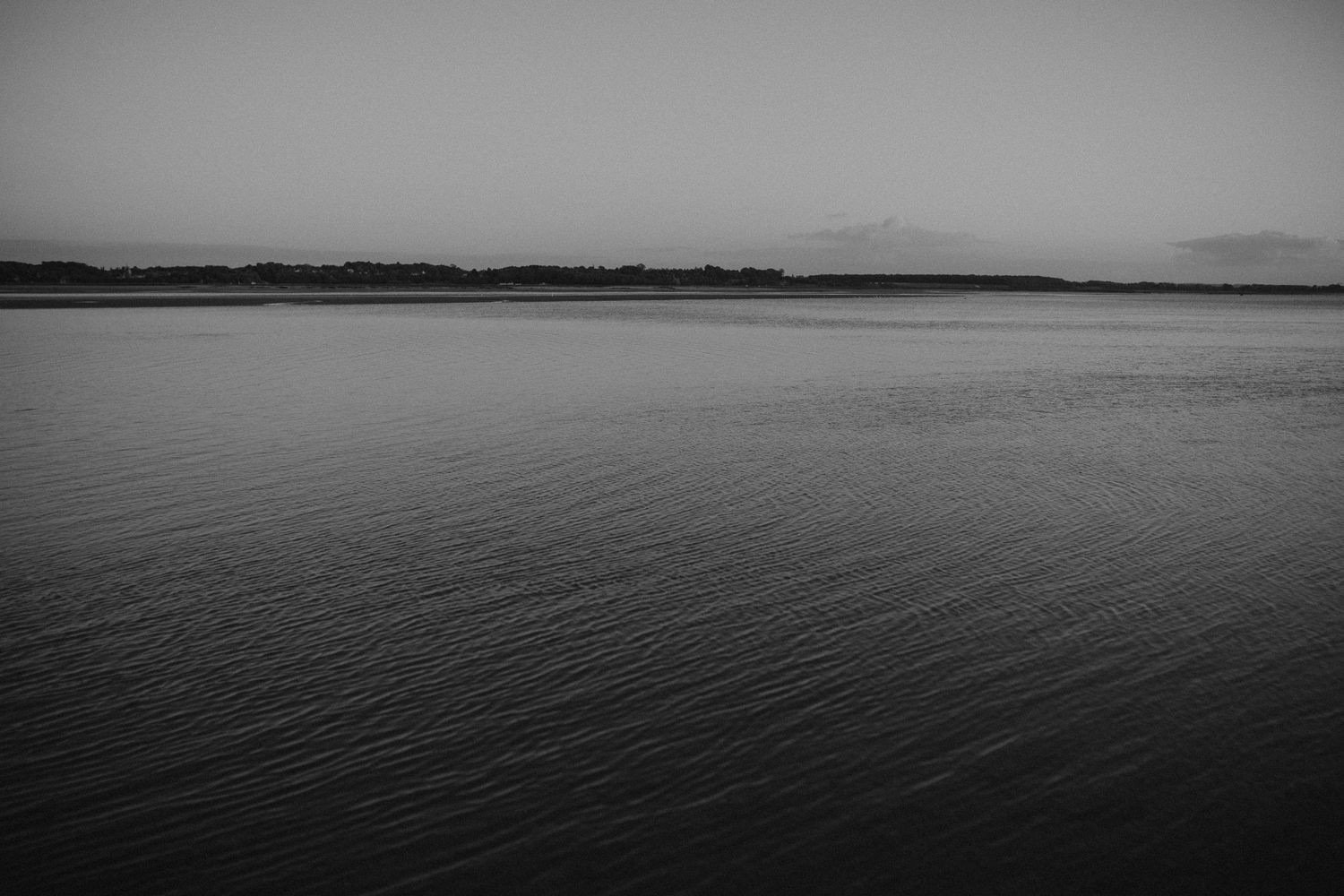 A serene black and white seascape with calm waters stretching to a distant horizon where mountains meet the sky.