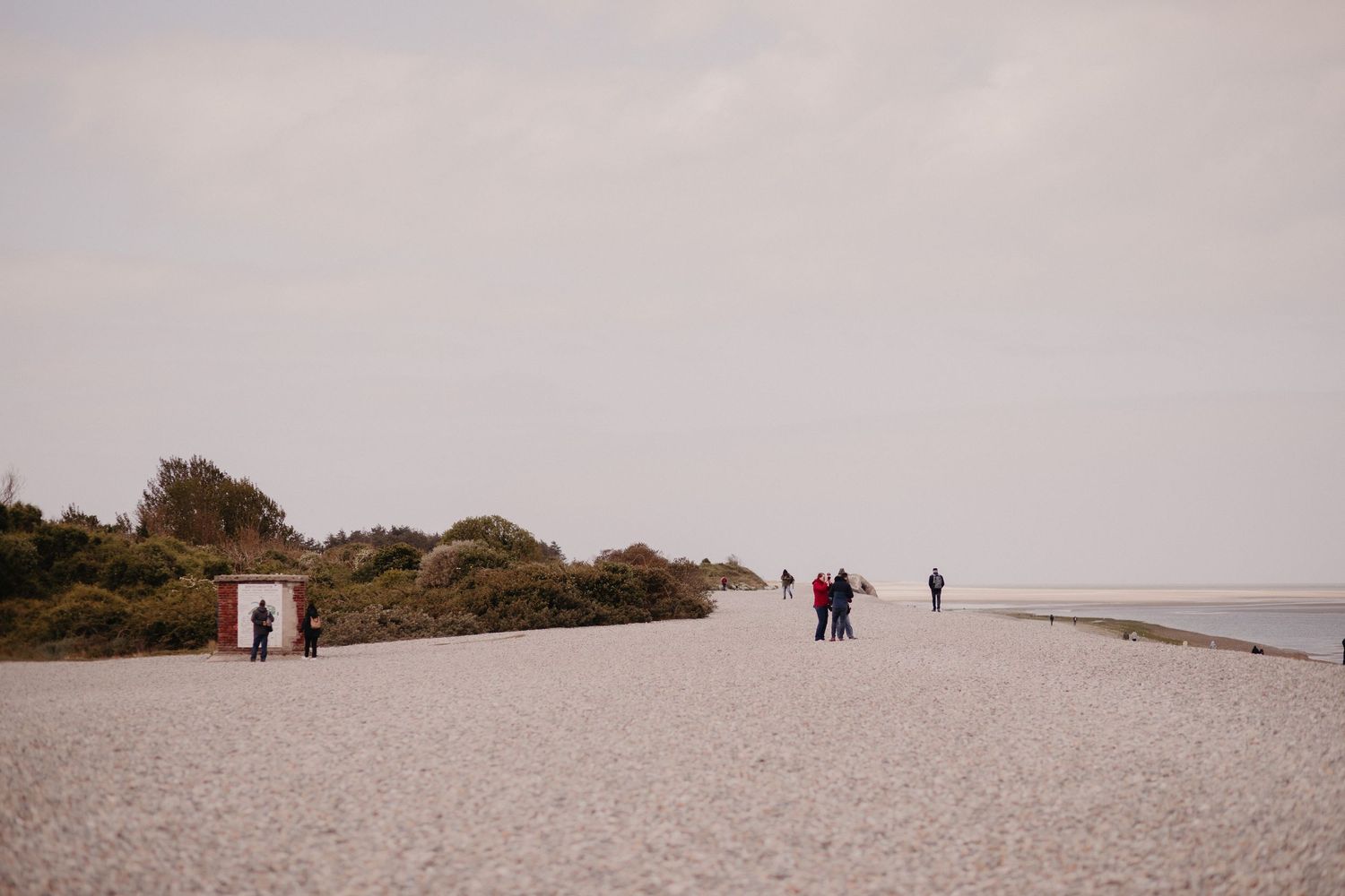 People walking along a misty beach with sand dunes and shrubs on a cloudy day at the coast.