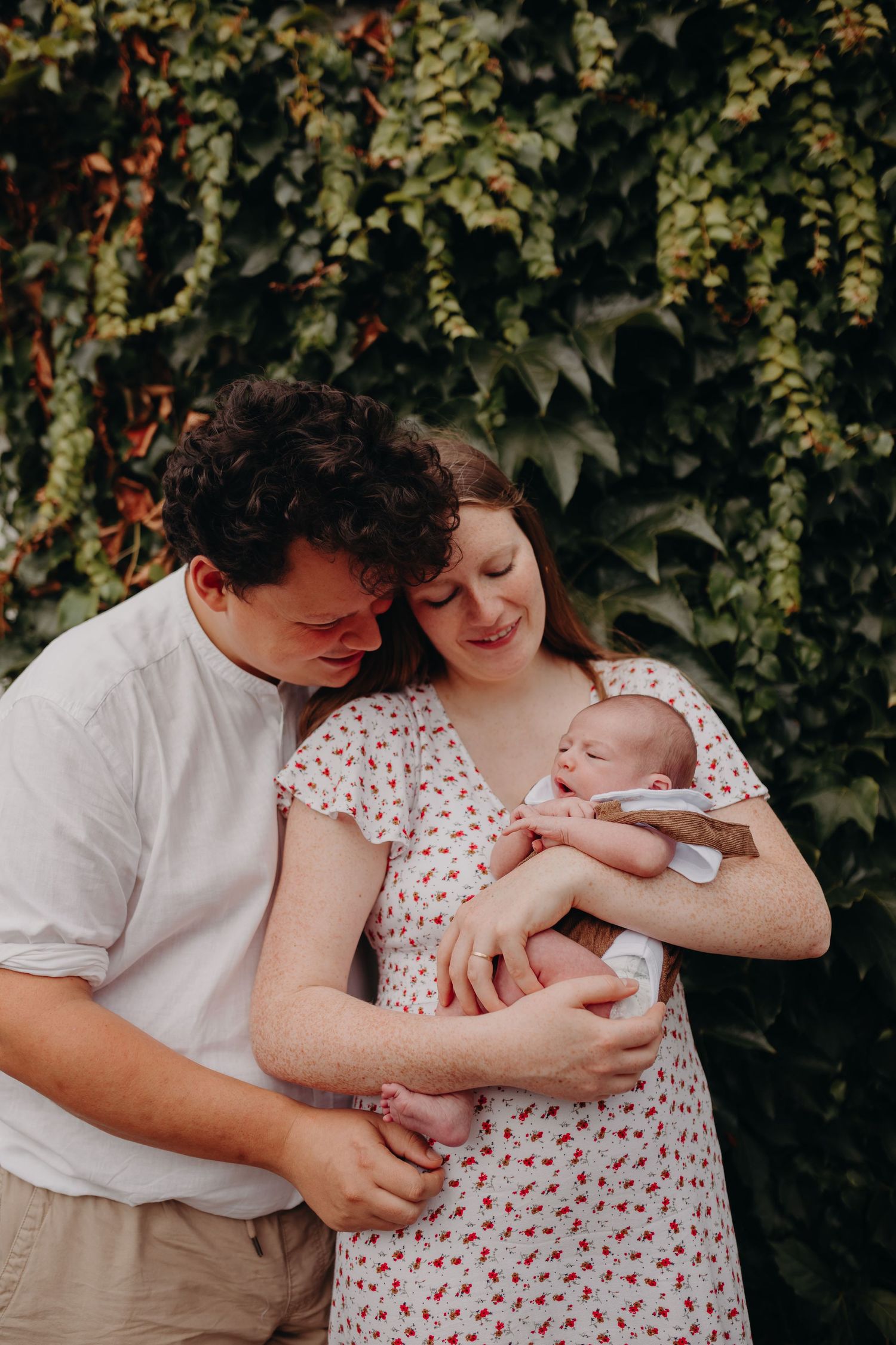 A couple holding their newborn baby while standing in front of a lush green plant wall in summer.