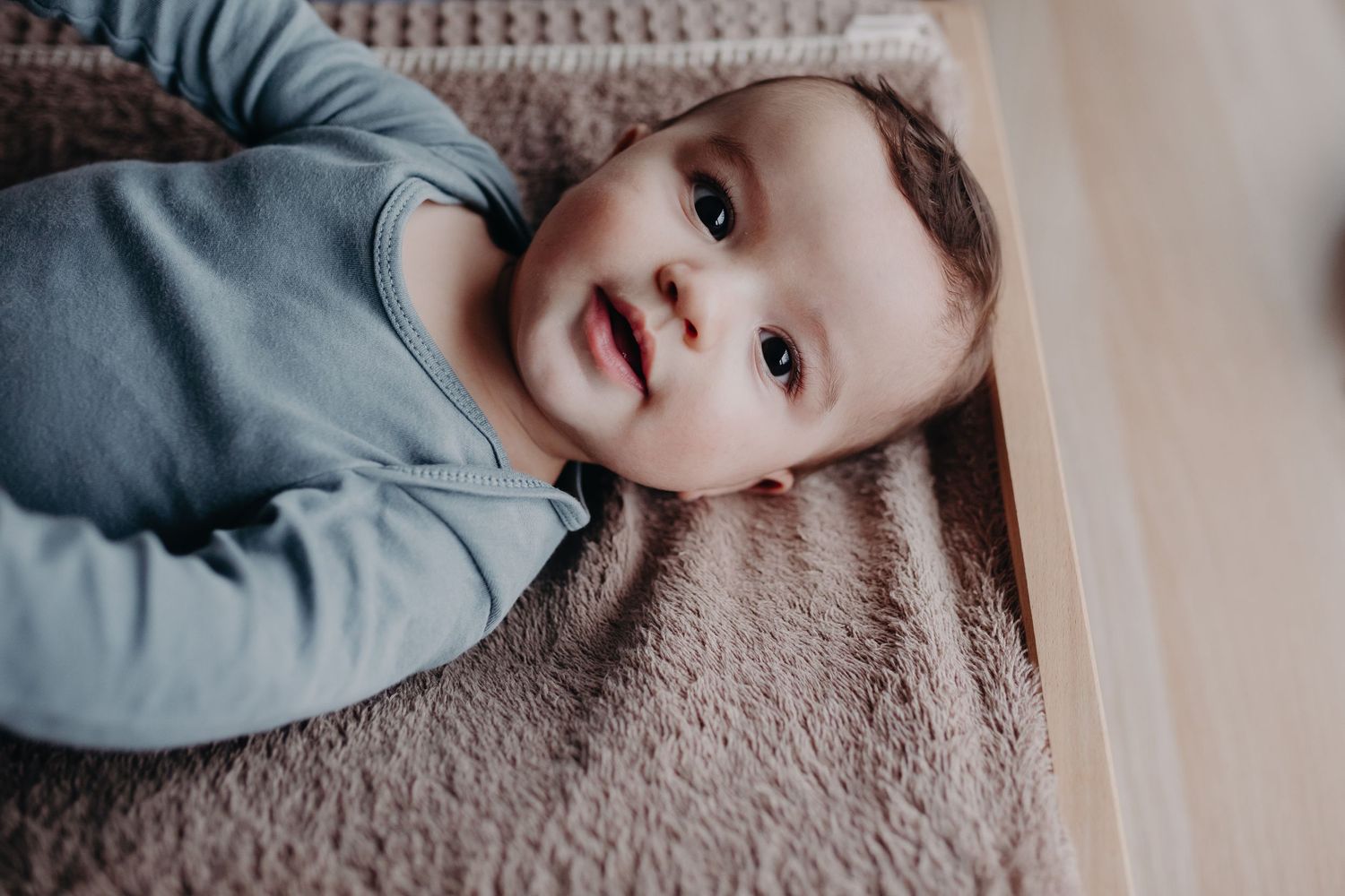 A smiling child wearing a blue-grey sweater lies on a plush beige carpet looking up at the camera.