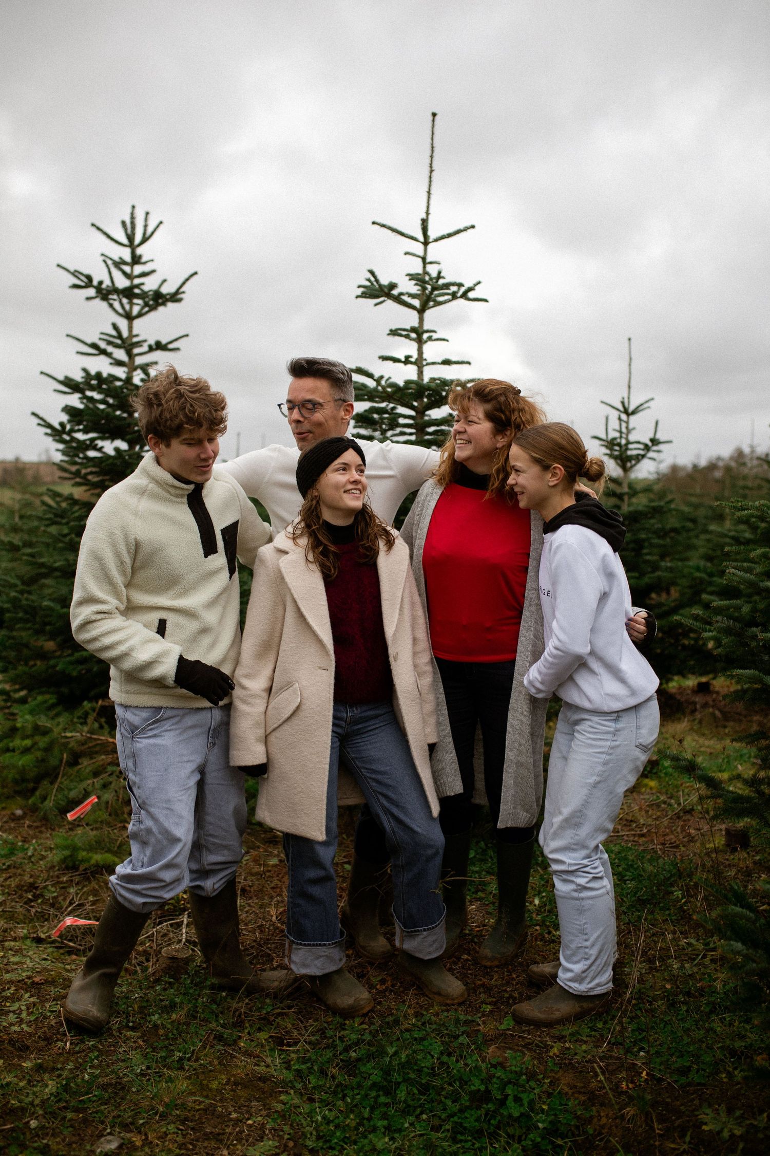 A group of friends in winter coats huddle together among evergreen trees on an overcast day.