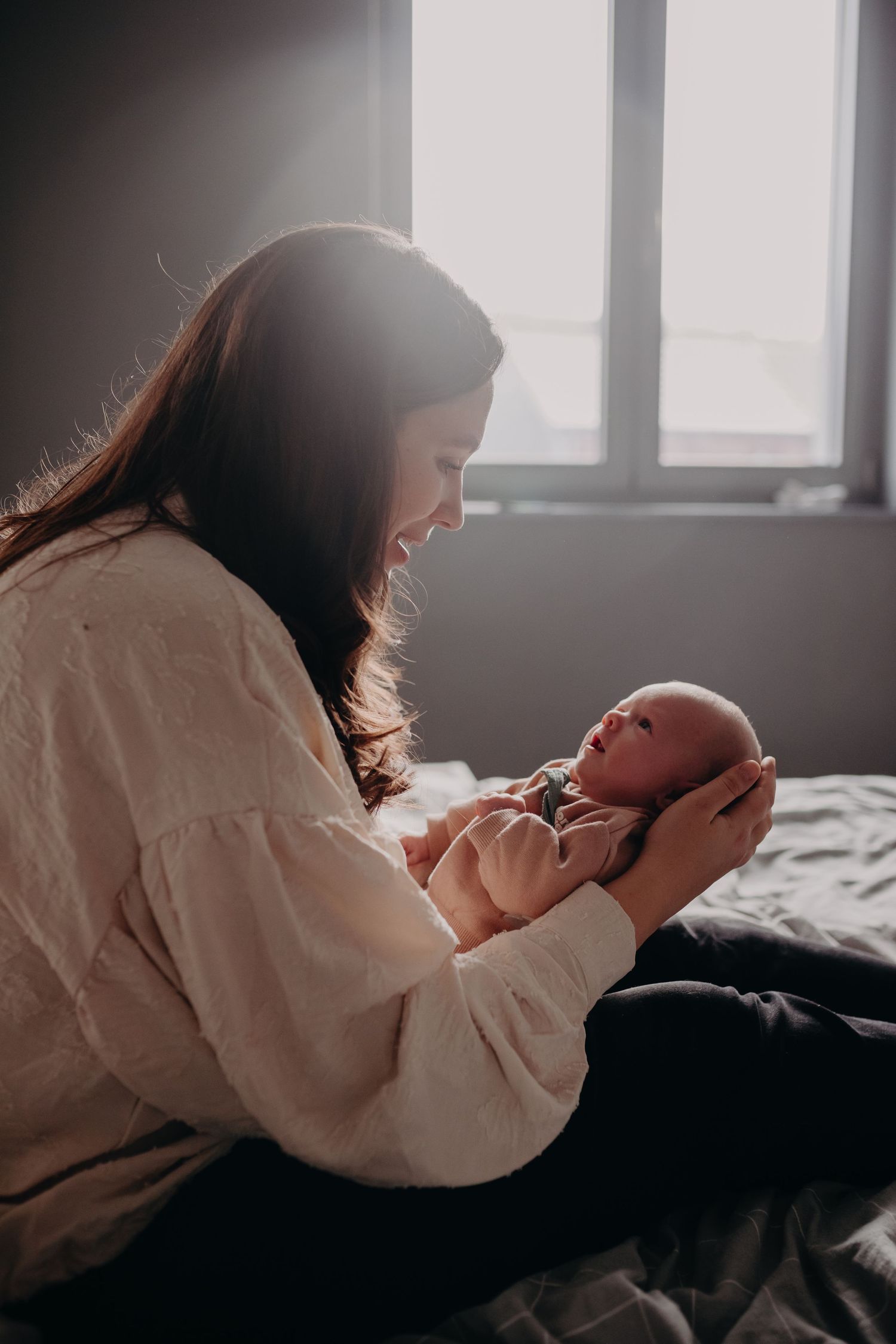 A person in a white sweater holds a baby while sitting on a bed in soft window light.