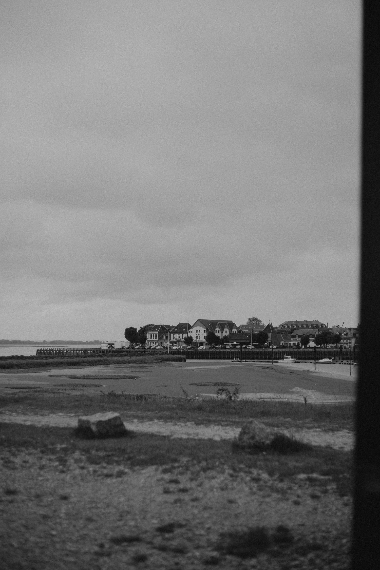 A black and white coastal landscape with buildings on the horizon and a moody overcast sky.