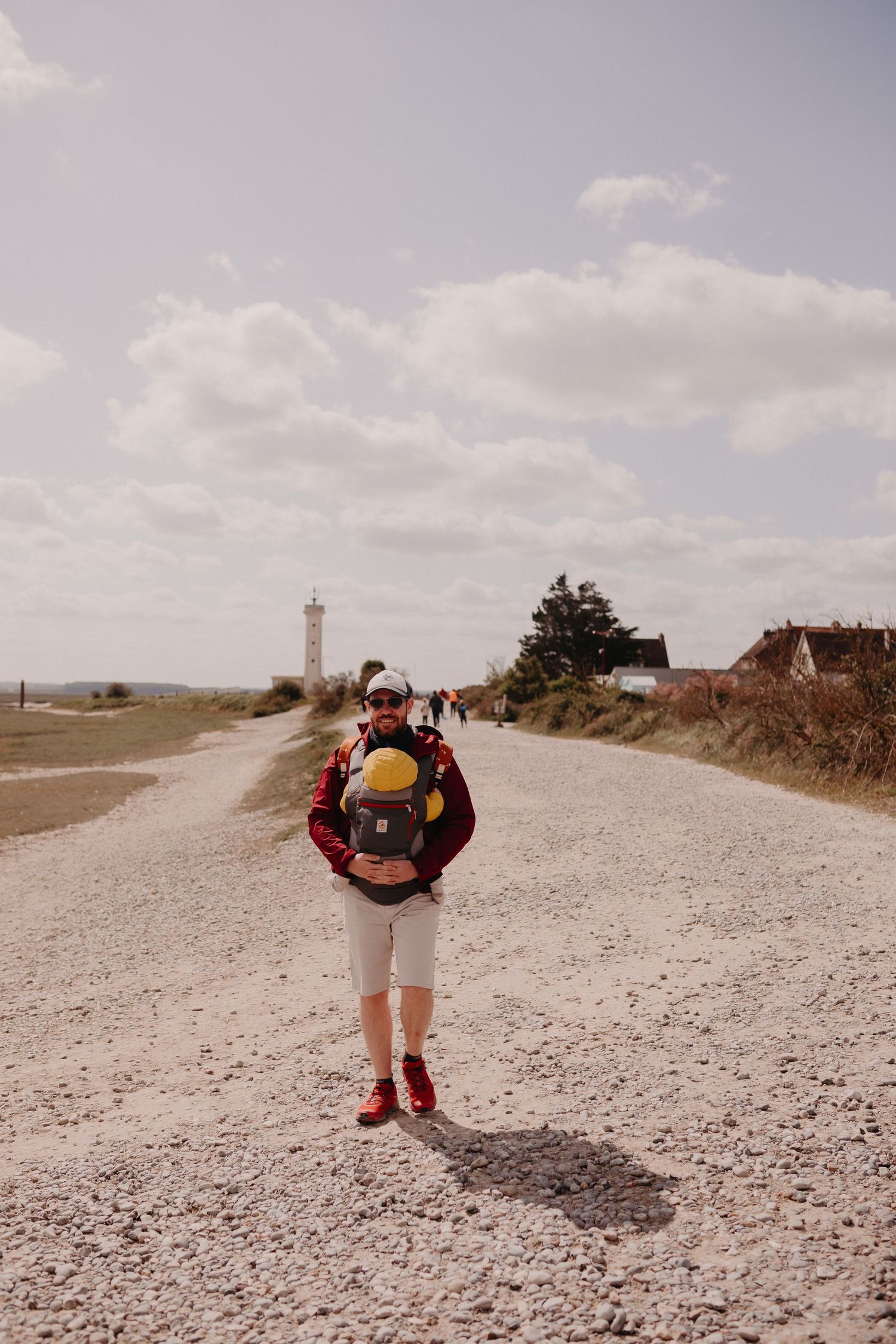 Person stands on a gravel path near a lighthouse with a backpack on a cloudy day.