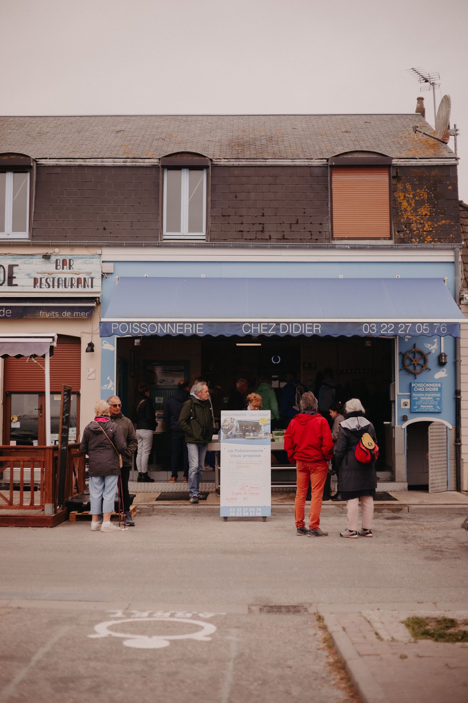 People waiting in line at a quaint seafood shop along a small town street with brick buildings and storefronts.