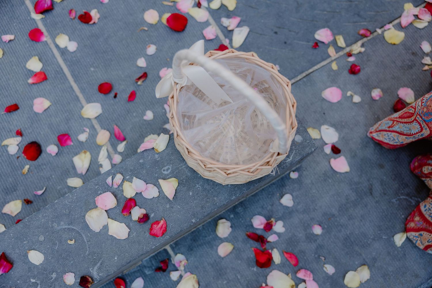 Flower girl basket with white ribbon surrounded by scattered rose petals on gray pavement at a wedding.